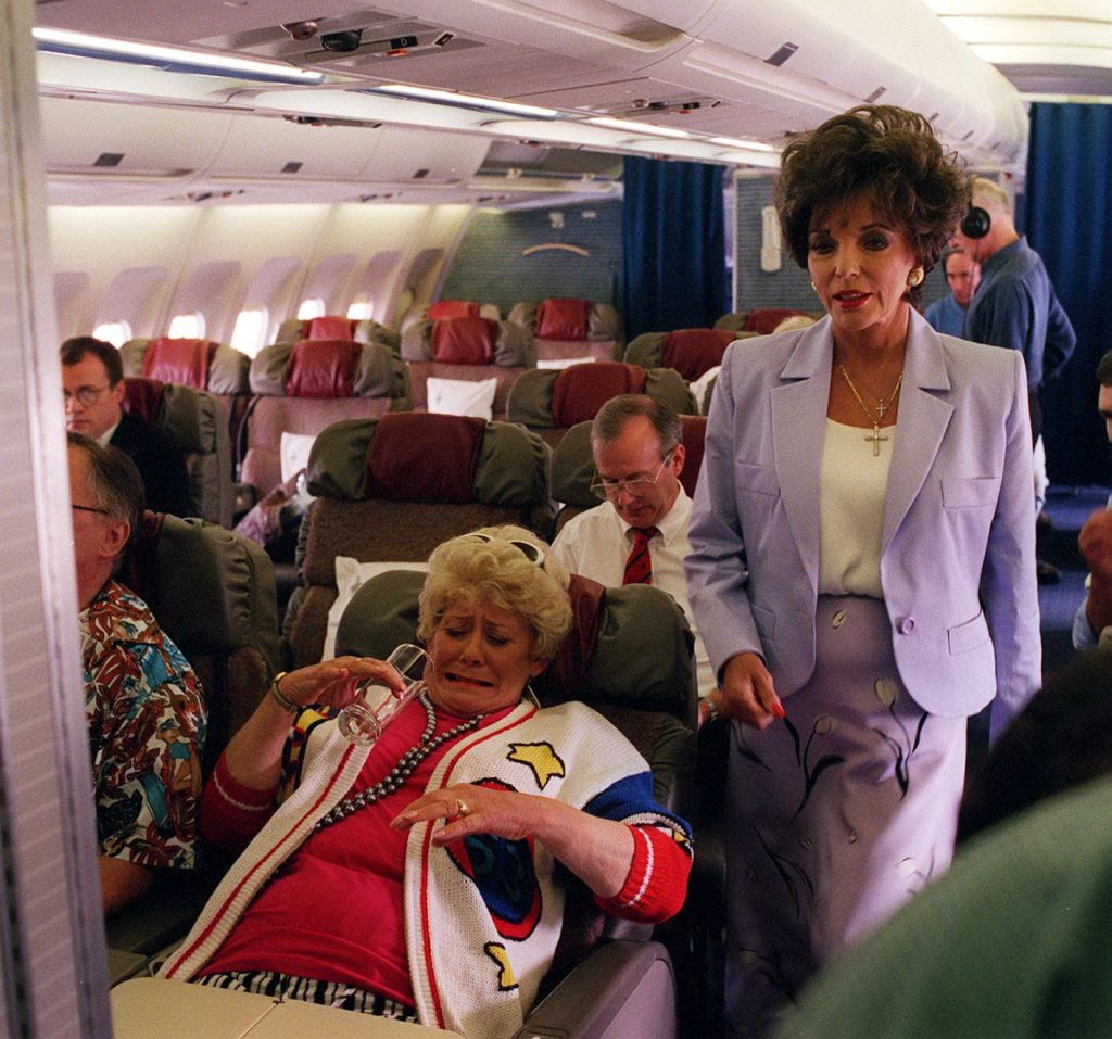 Joan Collins walks down the aisle of a plane as Vera Duckworth looks horrified to have spilled a drink on herself