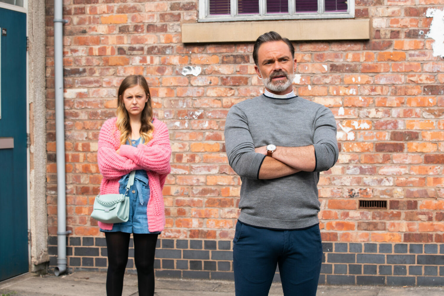 Coronation Street Summer and Billy look annoyed and defiant with their arms folded outside their flat