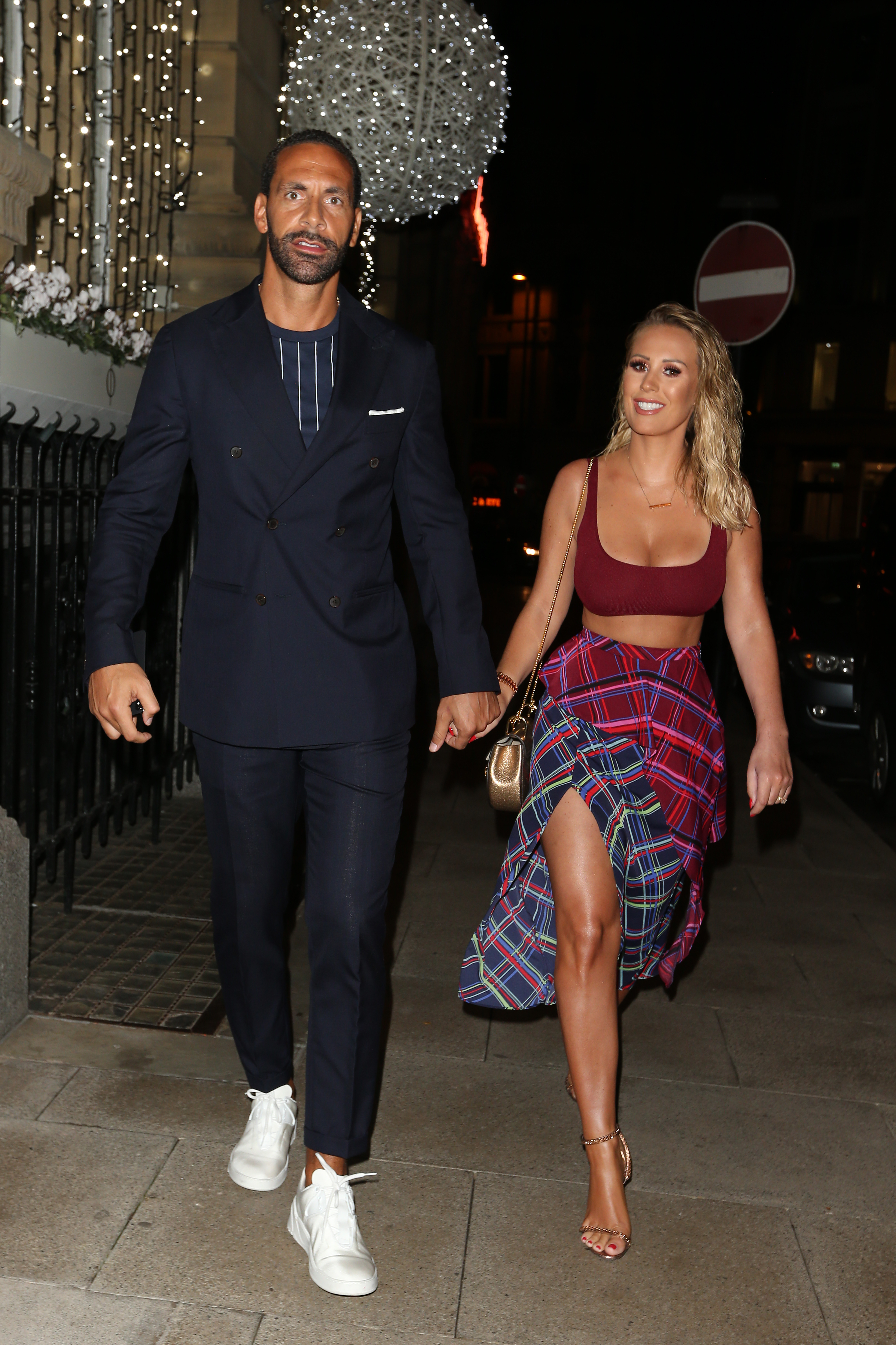 Rio Ferdinand in a black suit and grey t-short paired with white trainers and Kate Ferdinand wearing a burgundy crop top, heels and a check shirt around her waist holding hands