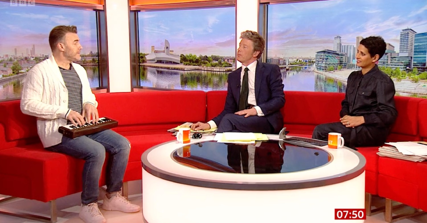 Gary Barlow sitting on a red sofa with his mini keyboard on his knees. Charlie Stayt and Naga Munchetty are sitting to the right of him on BBC Breakfast