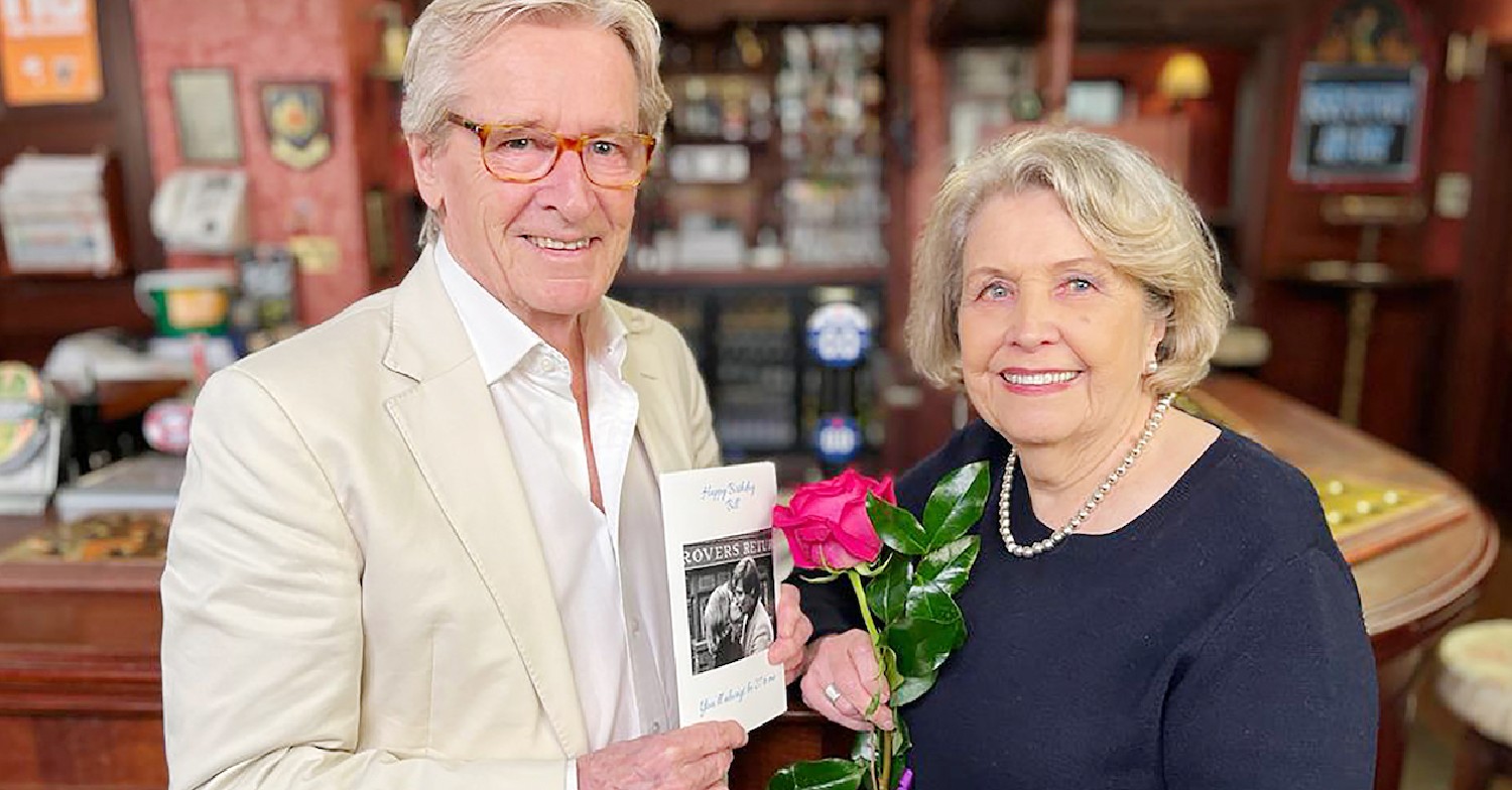 Anne Reaid and Bill Roach smiling at camera with a flower