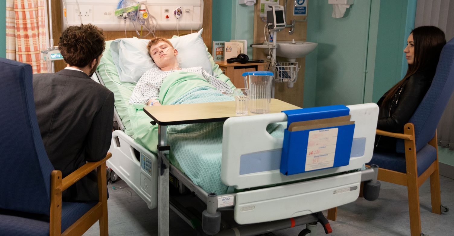 Max lies in a hospital bed after his operation with David and Shona in chairs in coronation Street