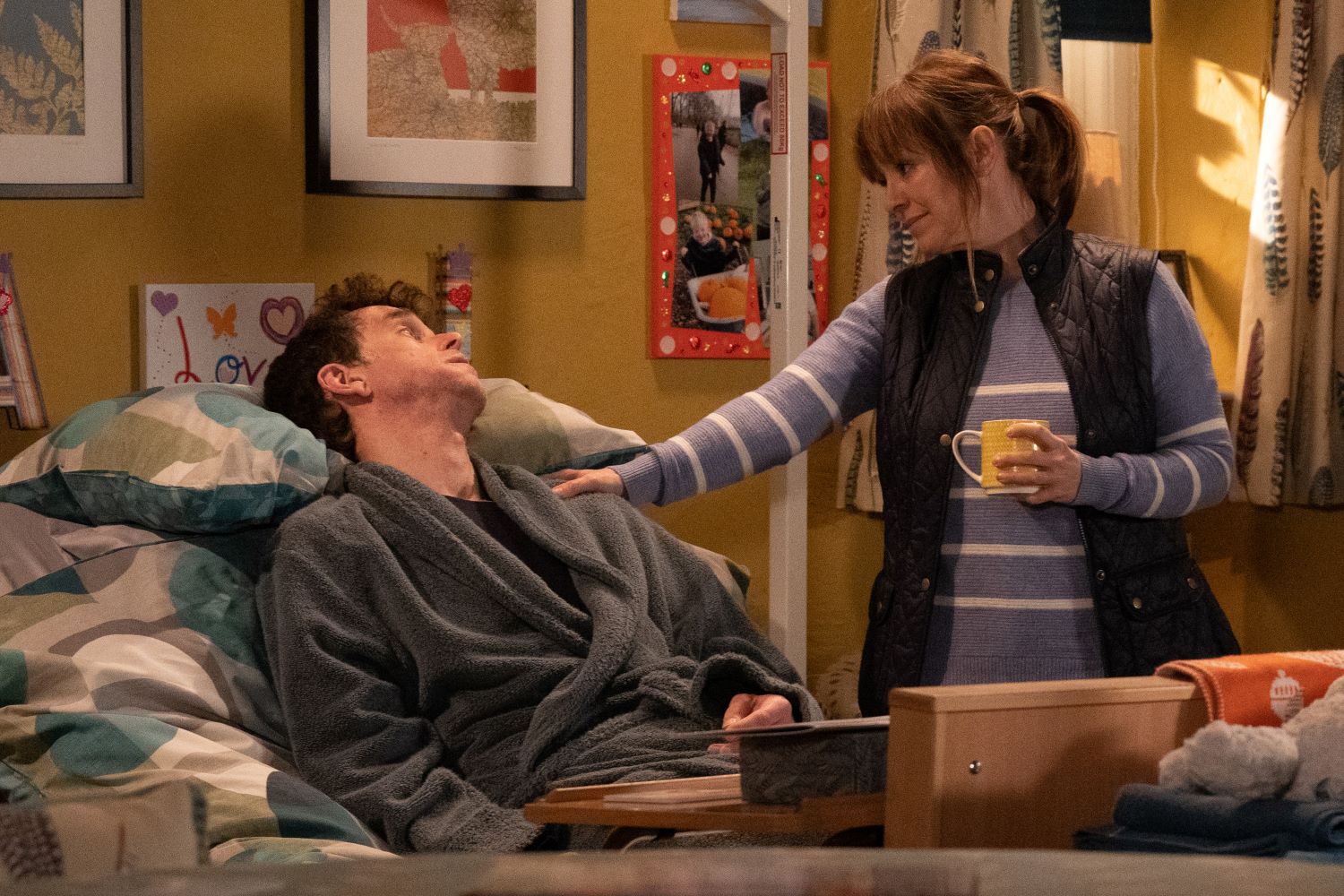 Marlon in bed as Rhona puts her hand on his shoulder holding her coffee