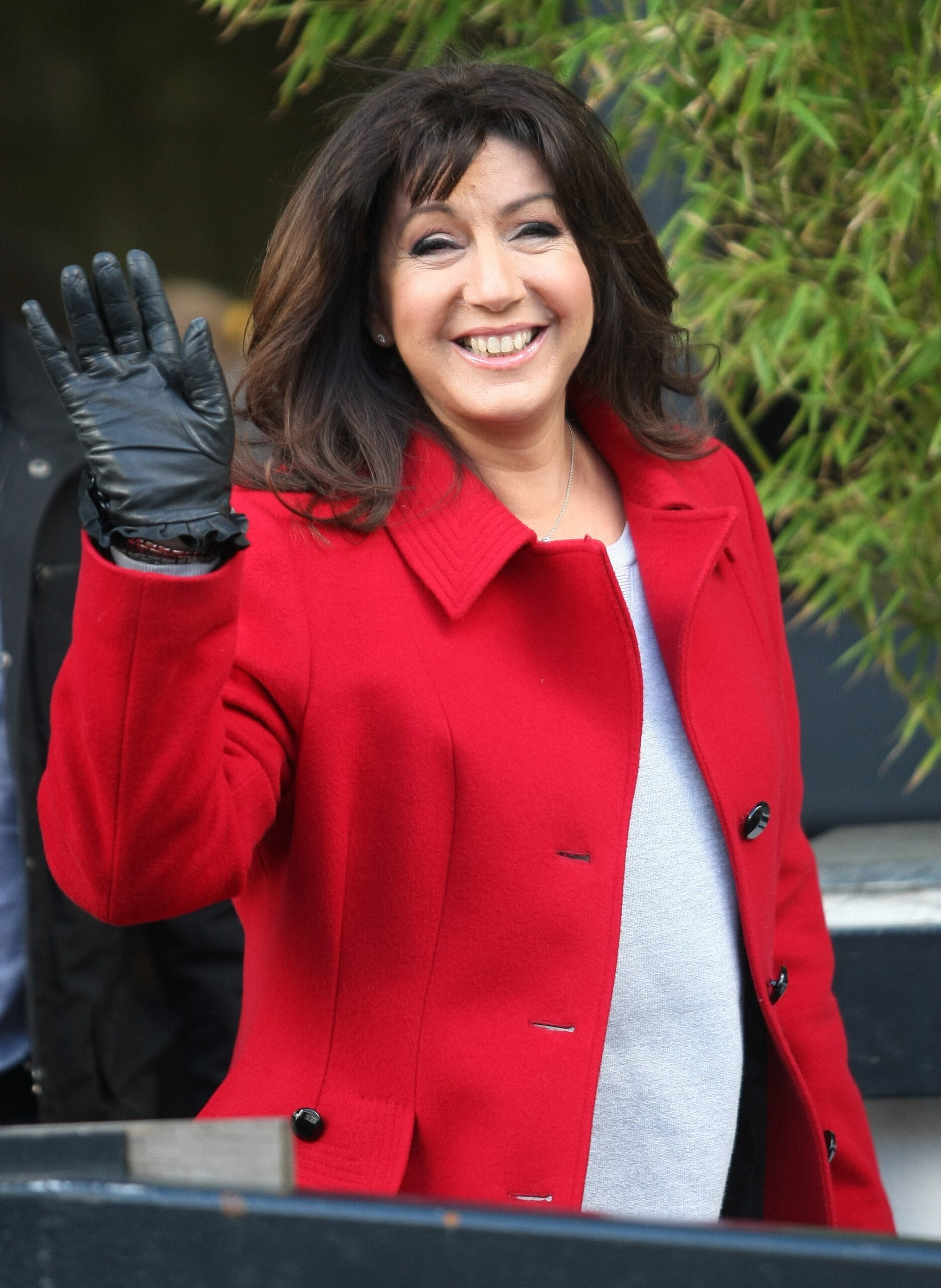 Jane McDonald smiling and waving wearing a read coat