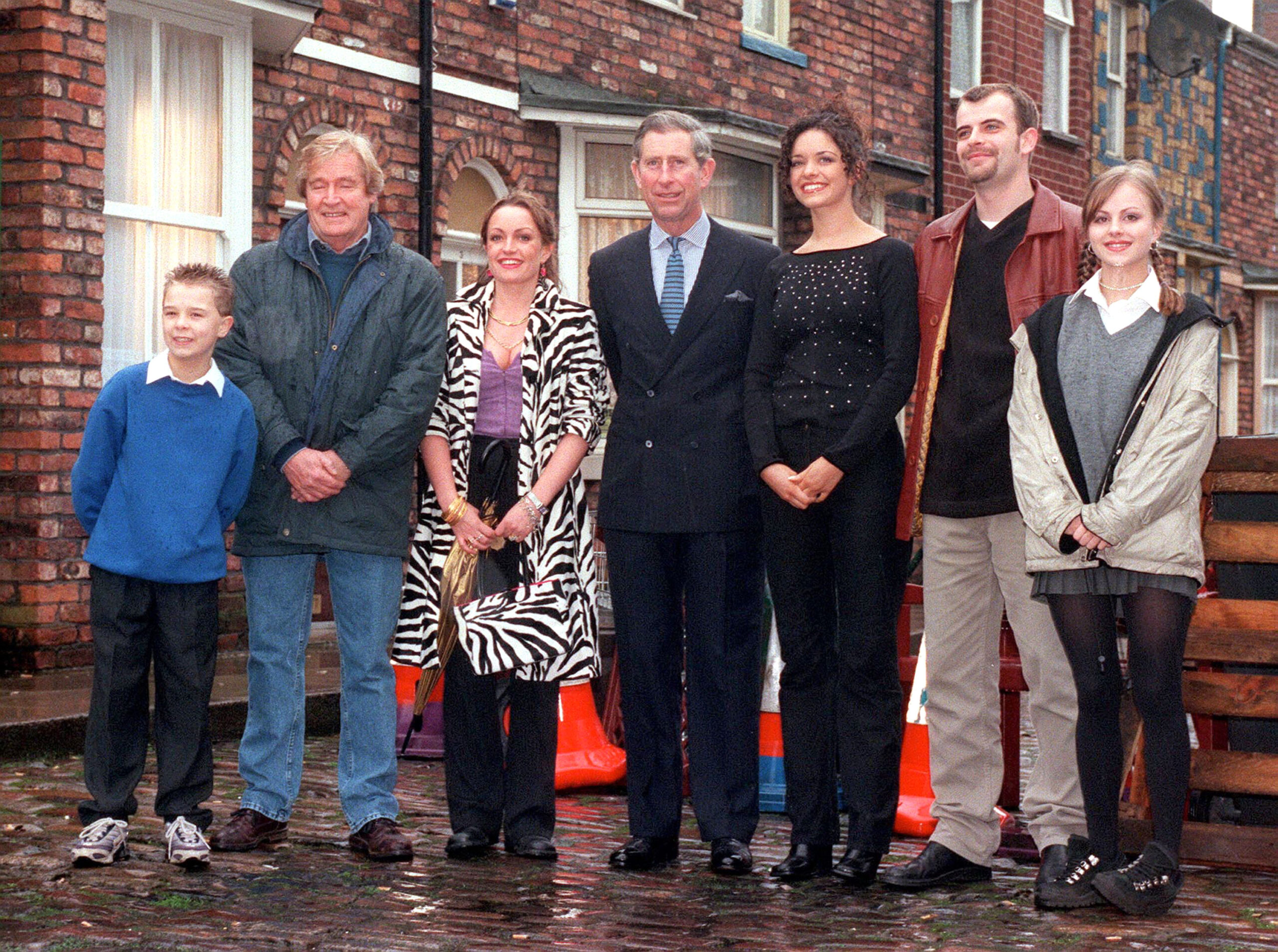 Prince Charles stands in the middle of Coronation Street cast members including Bill Roache, Jack P Shepherd and Simon Gregson on the rainy cobbles 