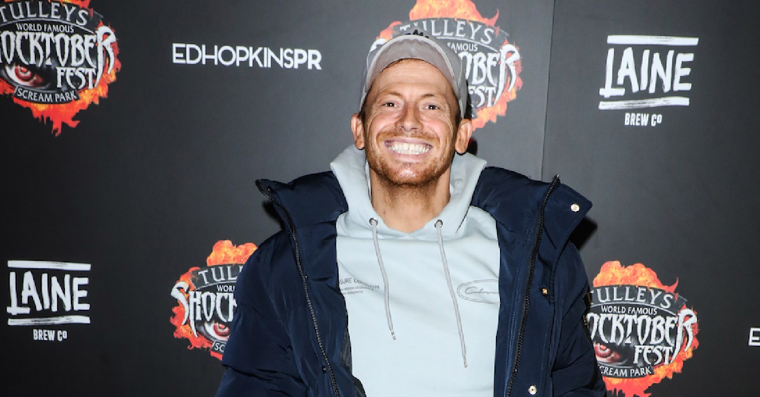 Joe Swash smiling wearing a grey hoodie, black jacket and baseball cap