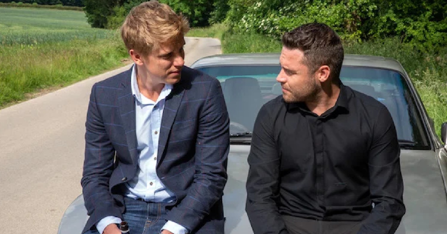 Emmerdale Robert and Aaron smile at each other while sitting on a car bonnet