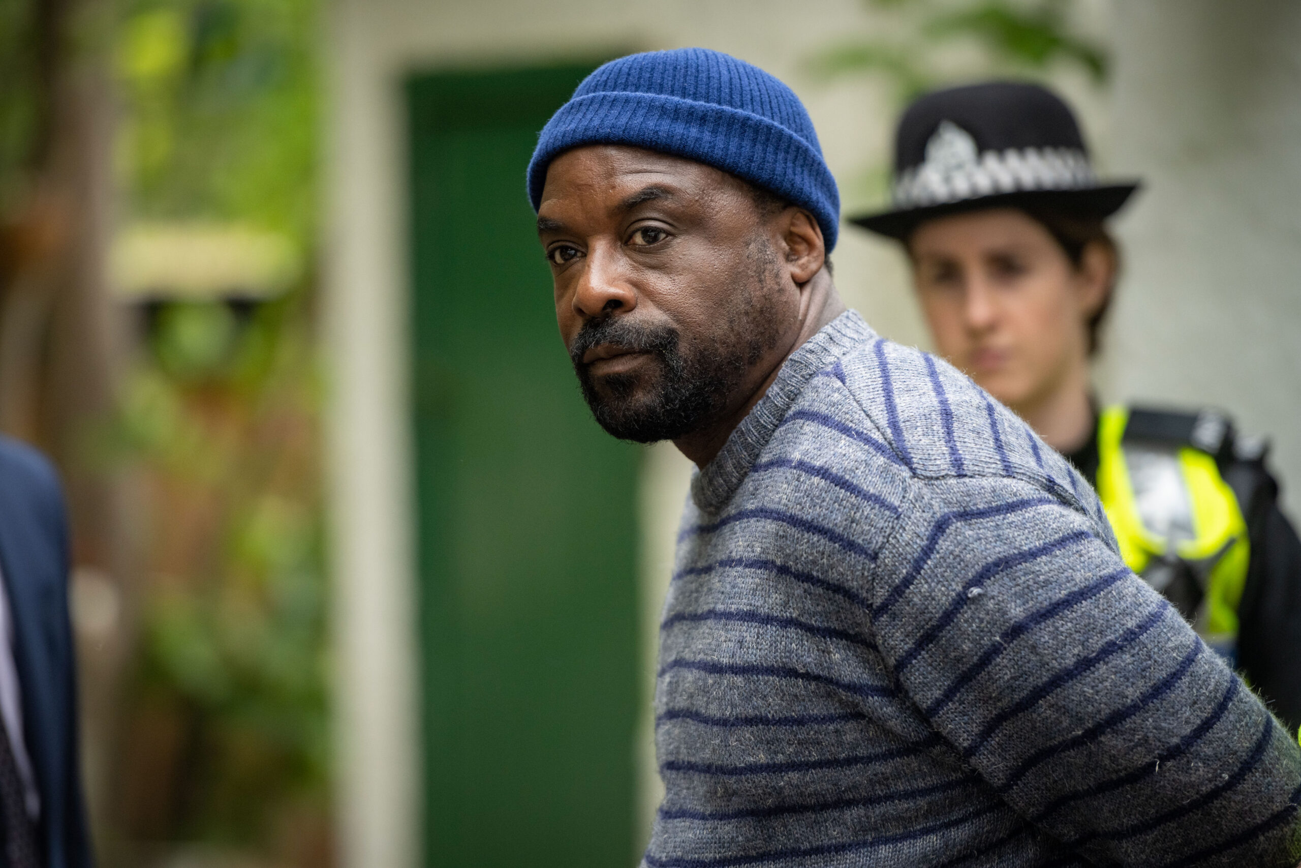 ARIYON BAKARE as Alex Snr., he stands with his arms in handcuffs outside his house with police next to him, frowning