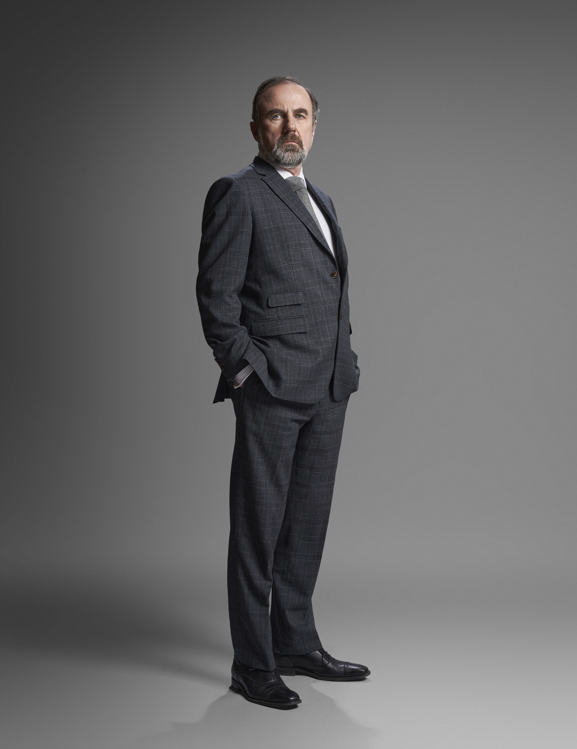 Bloodlands,Character Portraits,Jackie Twomey (LORCAN CRANITCH) He stands in a suit, with his hands in his pockets, he has a grumpy facial expression