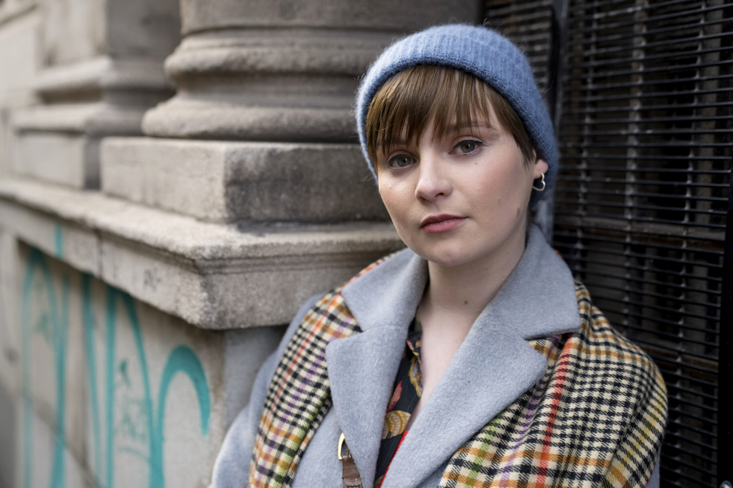 Bloodlands,Character Portraits,2,Izzy Brannick (LOLA PETTICREW) She stands outside in a city, she has a beanie on and she smiles