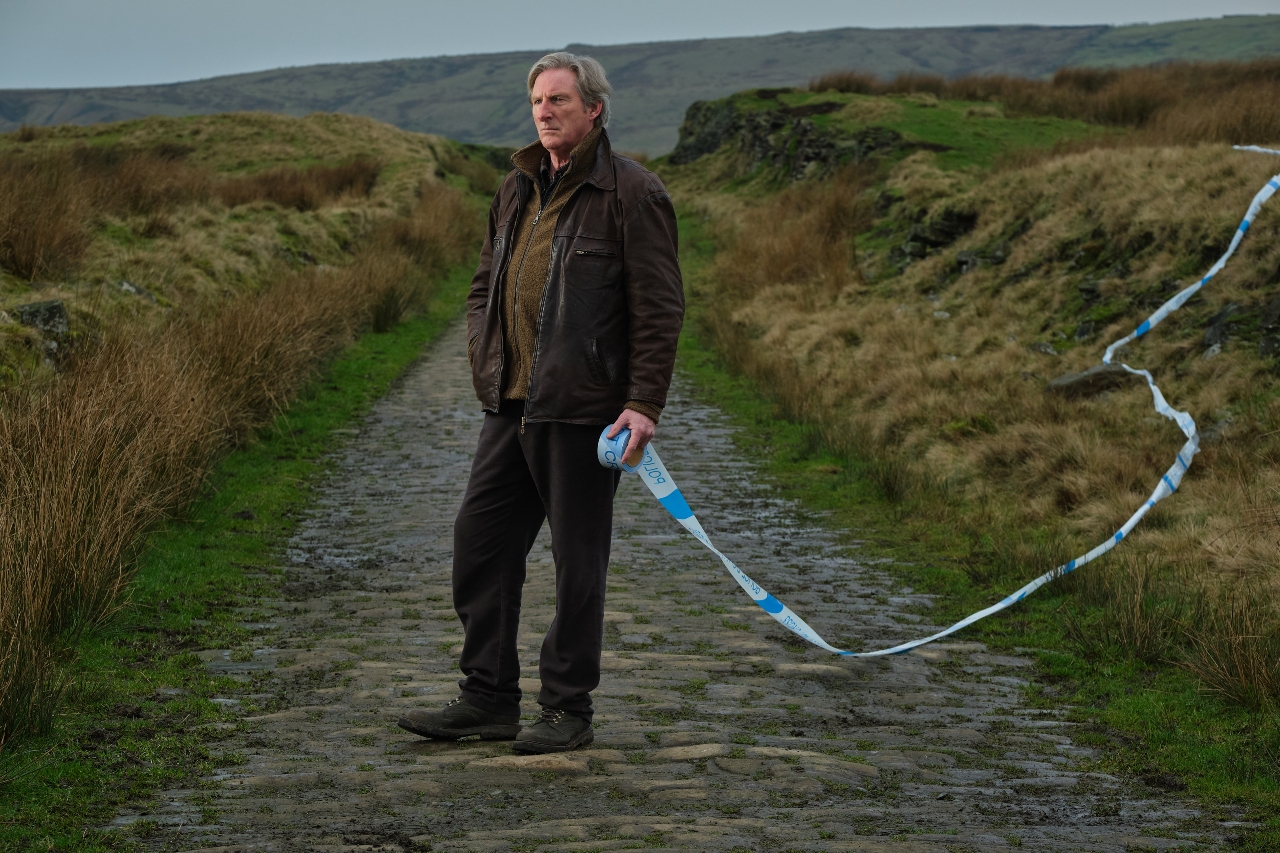 Adrian Dunbar as Alex Ridley, he stands in plain clothes in a field with police tape across the picture
