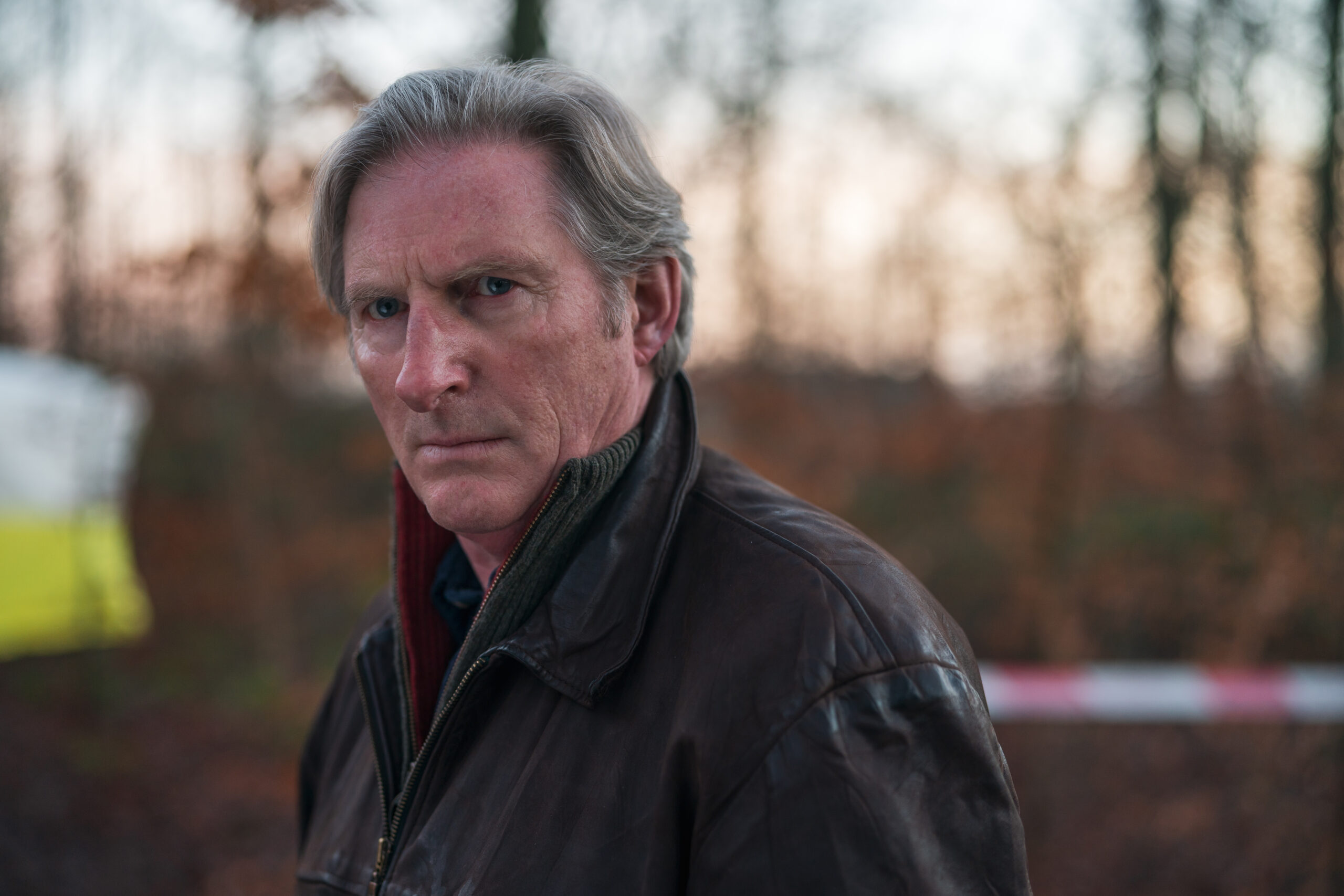 Adrian Dunbar as Alex Ridley, he looks into the camera outside with police tape behind him
