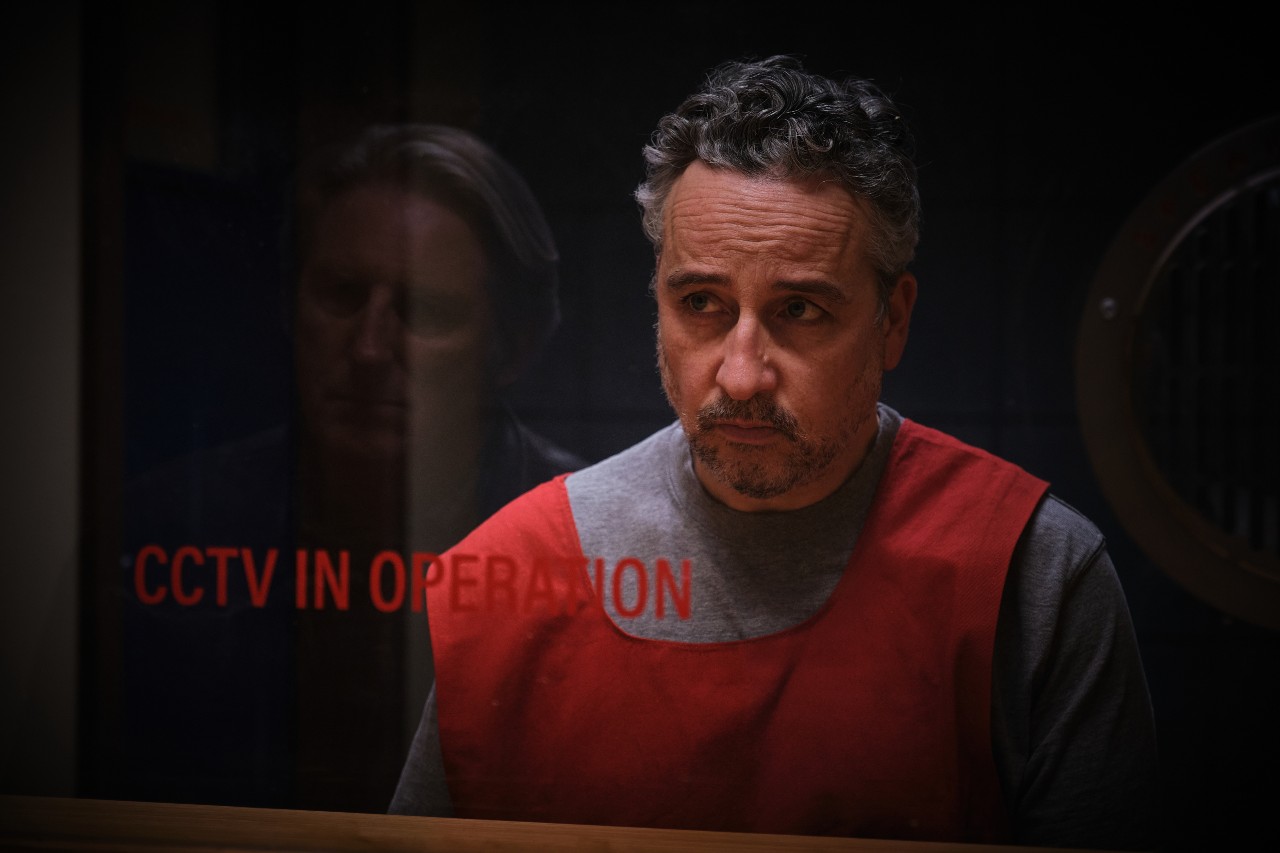 Aidan McArdle as Michael Flannery, he wears a red prison vest as he looks at Ridley in a prison visiting box