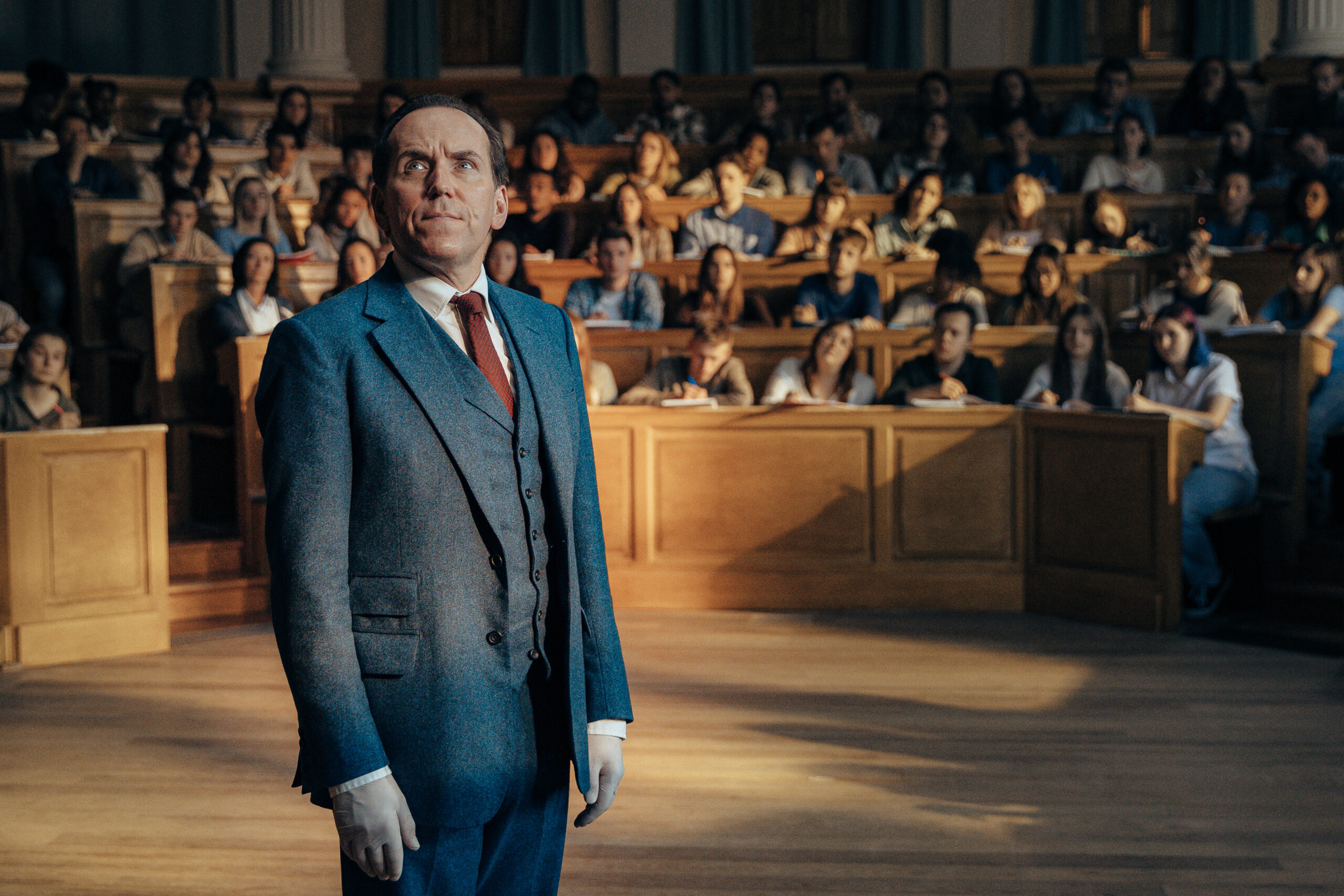Professor T stands in a lecture hall with a suit on and sad expression