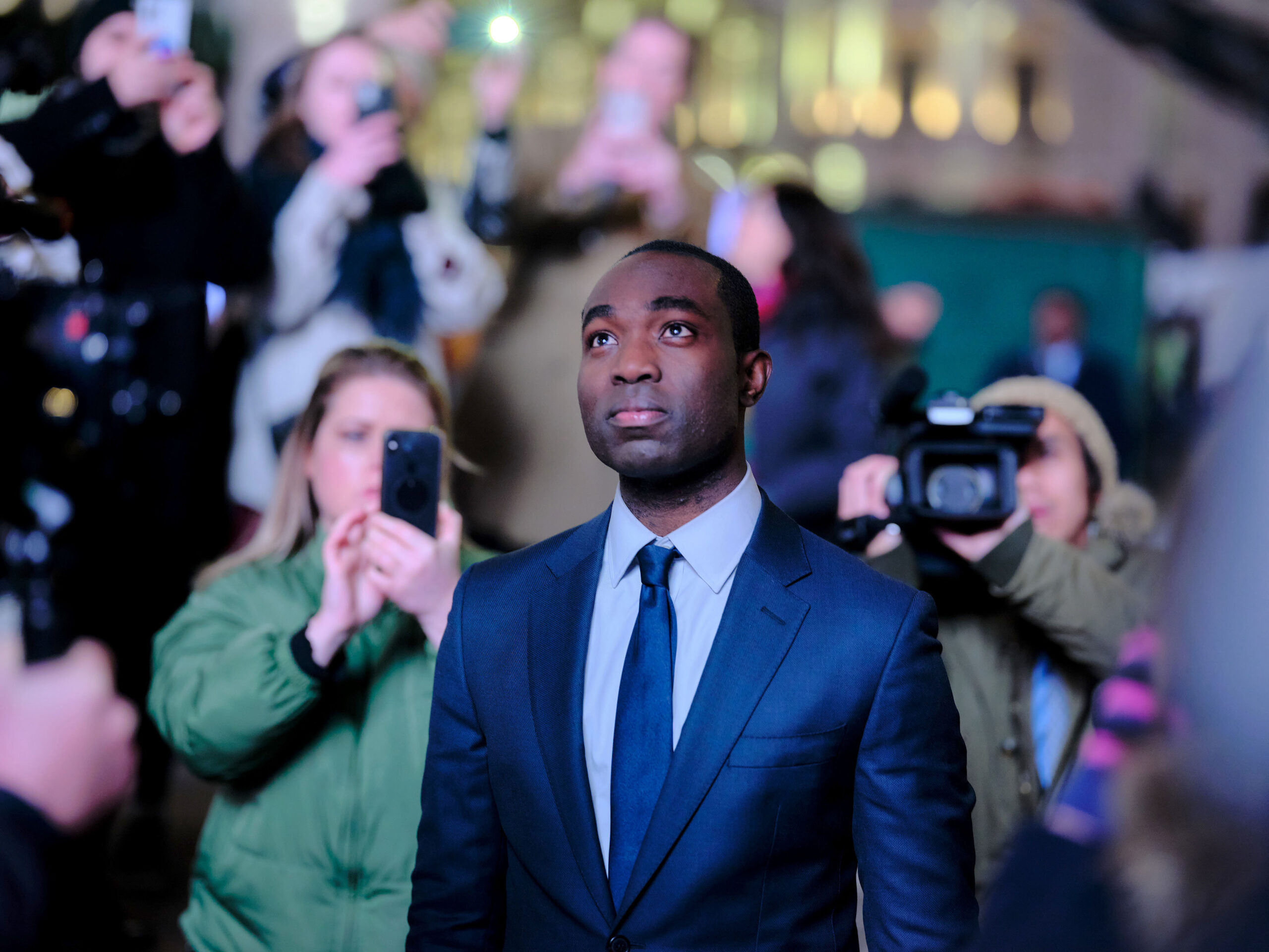 Paapa Essiedu as Issac Turner MP, he is a crowd with cameras and mobile phones as he stares up at a screen, in shock
