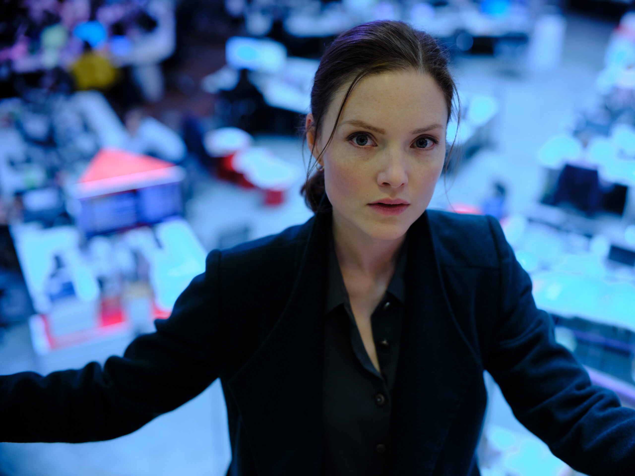 Holliday Grainger as Rachel Carney, she looks up the camera in a black jacket, she's in a blue IT room with computers