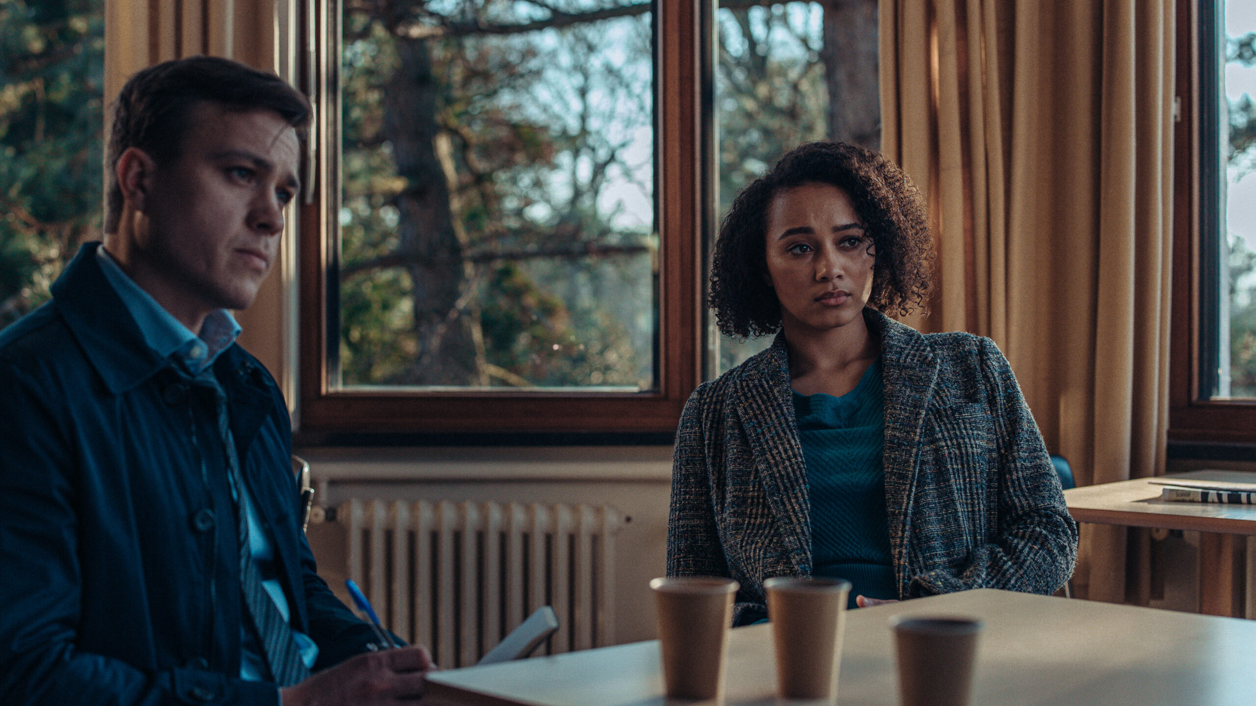 Barney White as Dan Winters (left) and Emma Naomi as Lisa Donkers in Professor T, they sit as a table with coffees in front of them as they interrogate a student