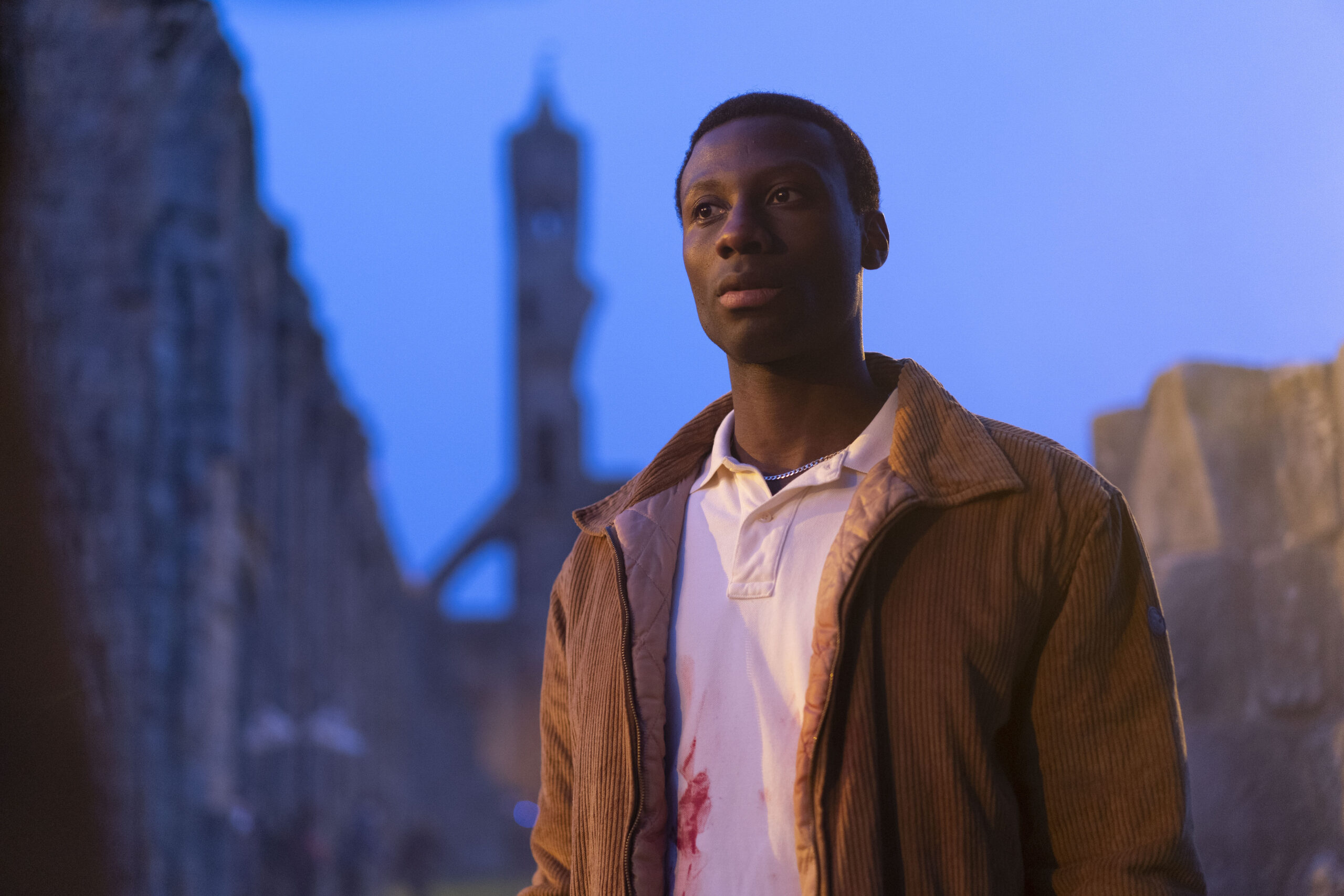 Boum Thingang as Alex, he stands in a dark street in a brown jacket