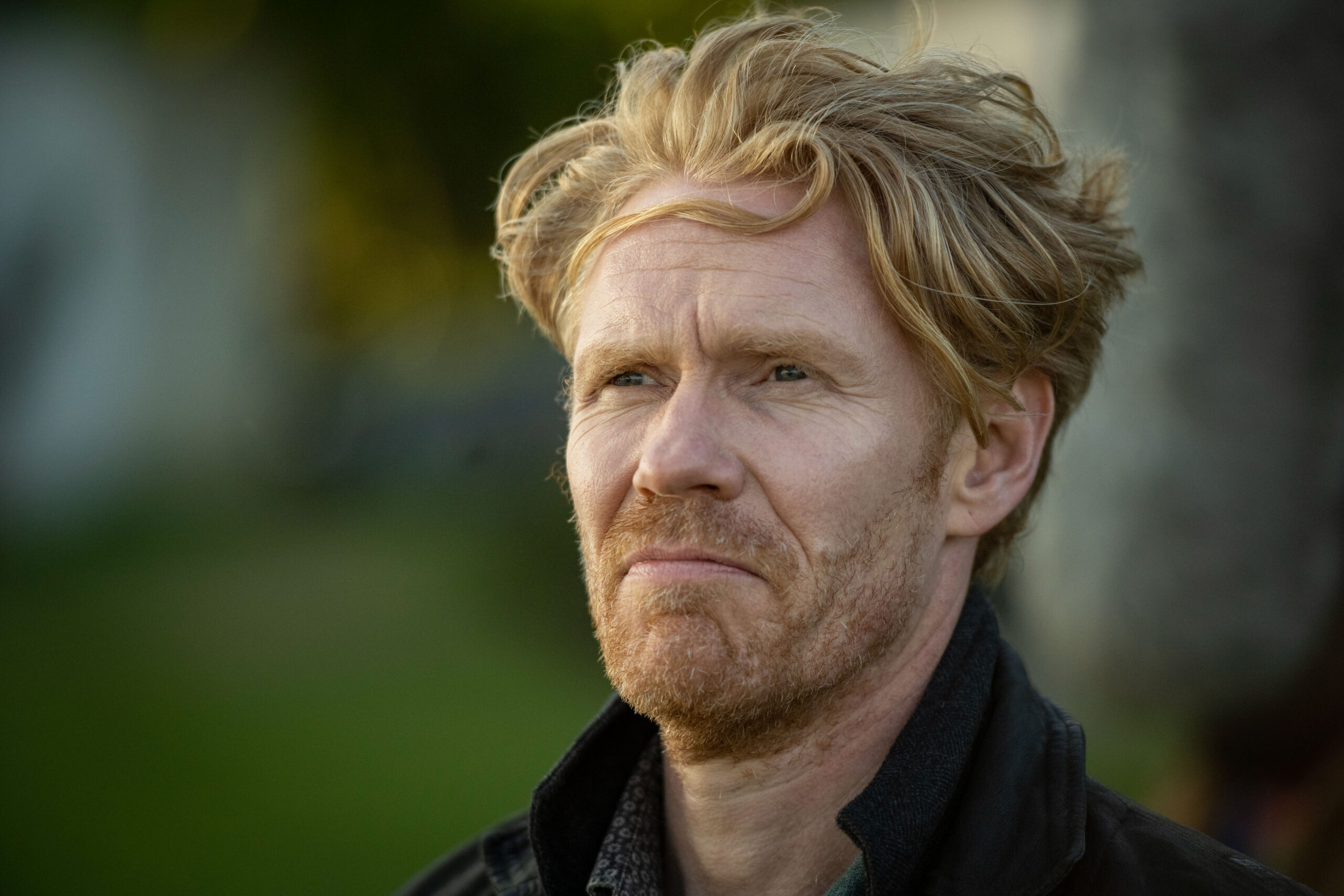 Michael Schaeffer as Tom Weird Sr, he stands in a park with a black shirt on looking upset