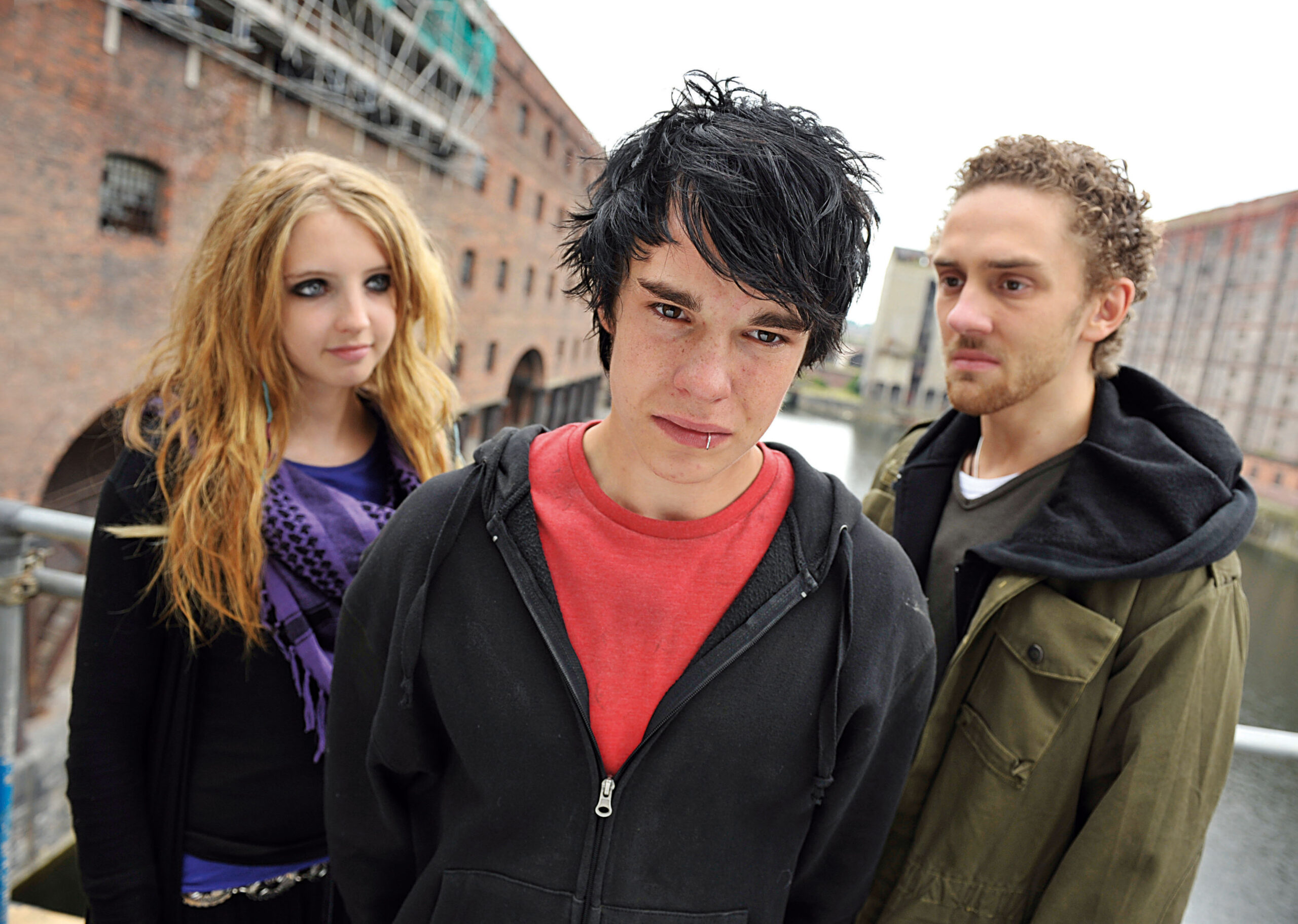 Nico Mirallegro as Newt in Hollyoaks, he stands in a group of people staring at the camera, he has a hoodie on and dark black hair