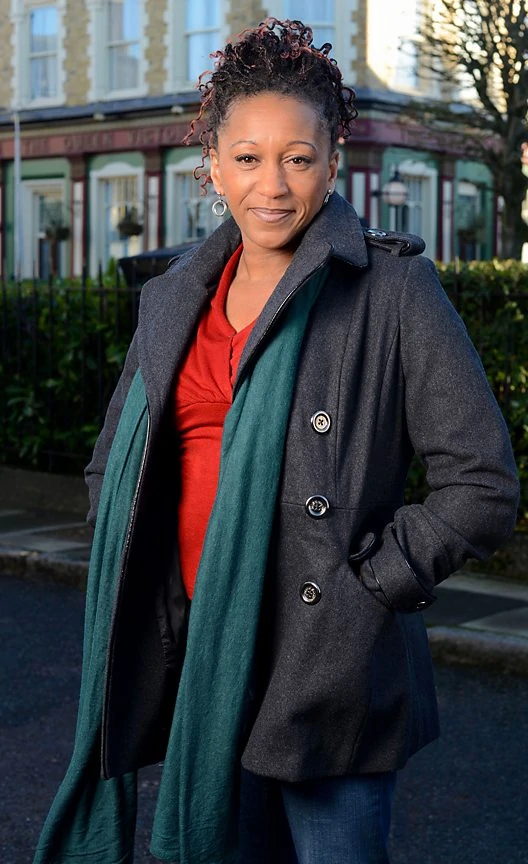 Clare Perkins as Ava in EastEnders, she poses in Albert Square with her hands in her pocket and a smile