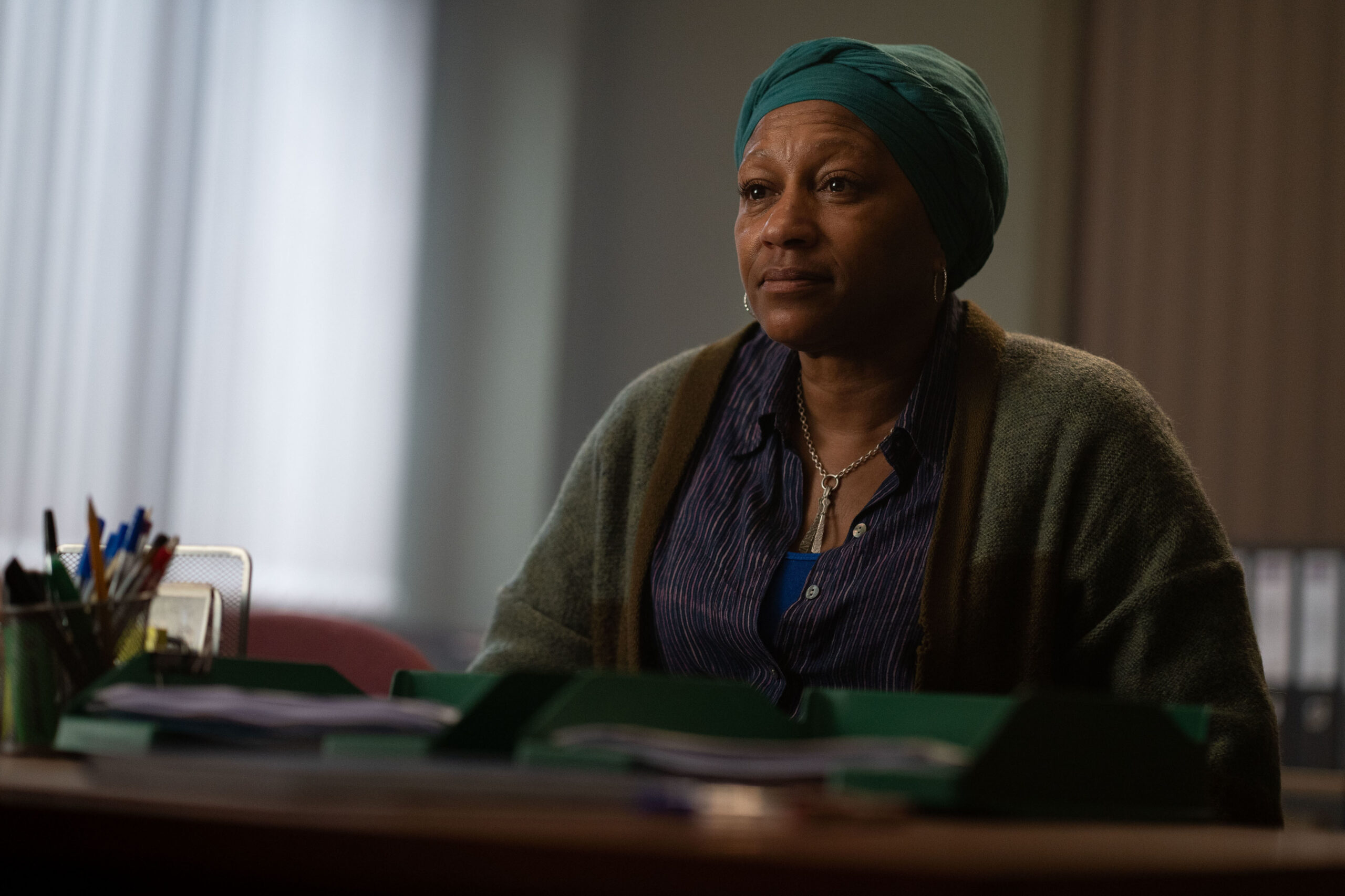 Clare Perkins as Maya Okeke in THE OUTLAWS, she sits at a desk with a neutral expression