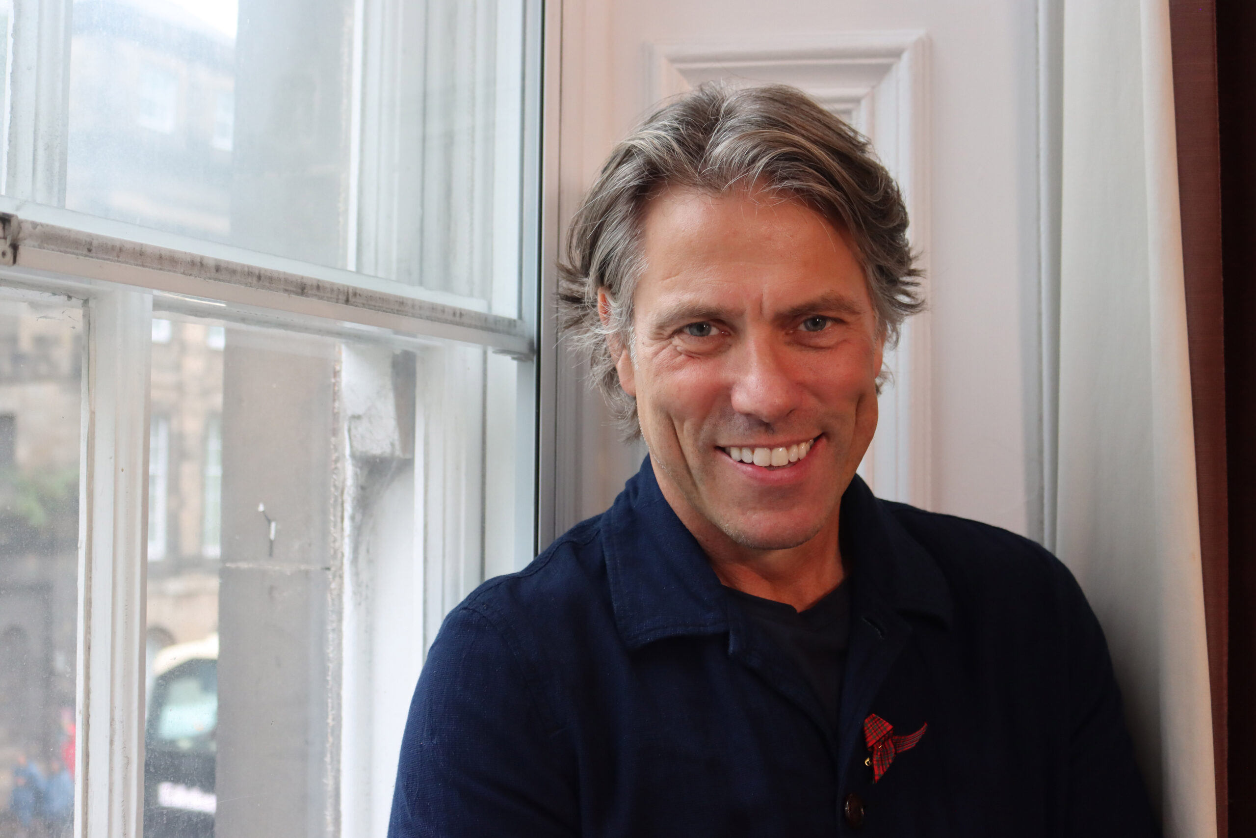 John Bishop in Life After Deaf, he smiles in a black shirt, leaning against a window
