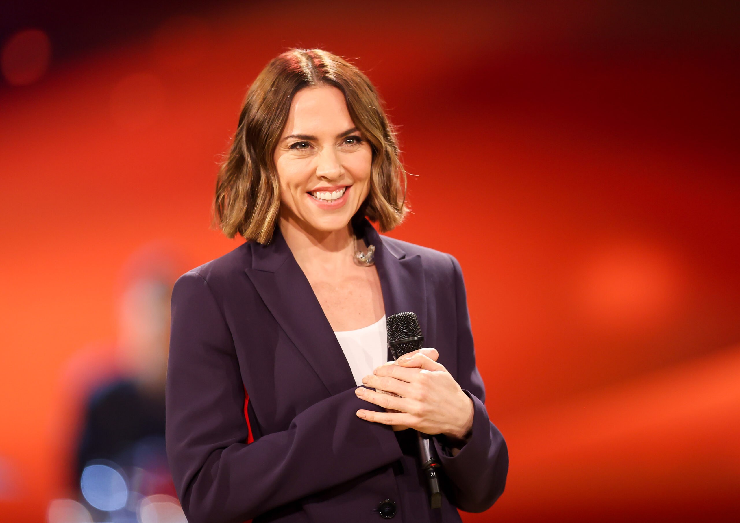 Mel C smiling in a black suit. Red background.