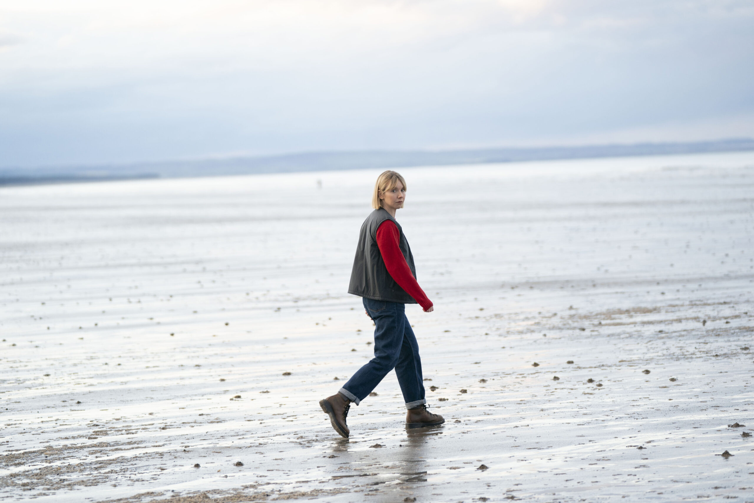 Lauren Lyle as Karen Pirie, she stands in a beach with a red and brown jacket on, looking back at the camera