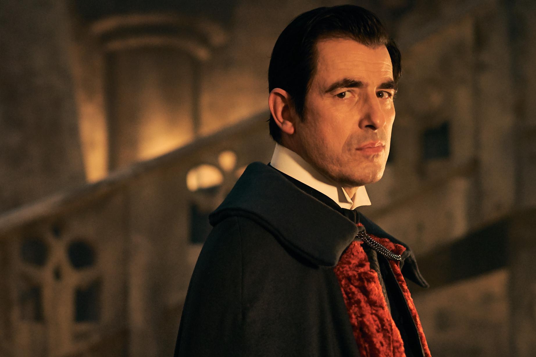 Claes Bang as Dracula in Steven Moffat show Dracula, he smiles slightly looking into the camera in a red and black vampire outfit