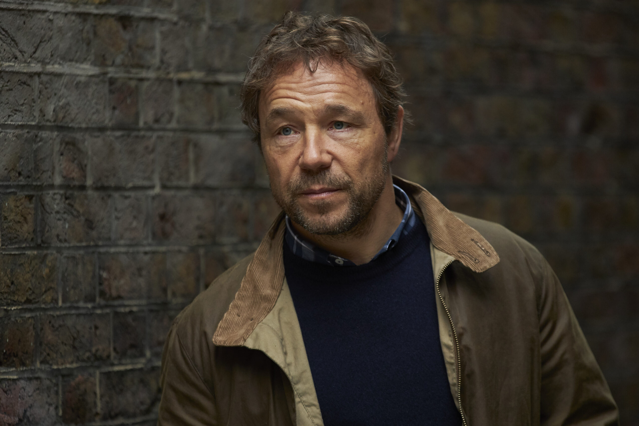 Stephen Graham as Matthew Collins in The Walk-In, he stands against a wall in a brown jacket and has a small smile