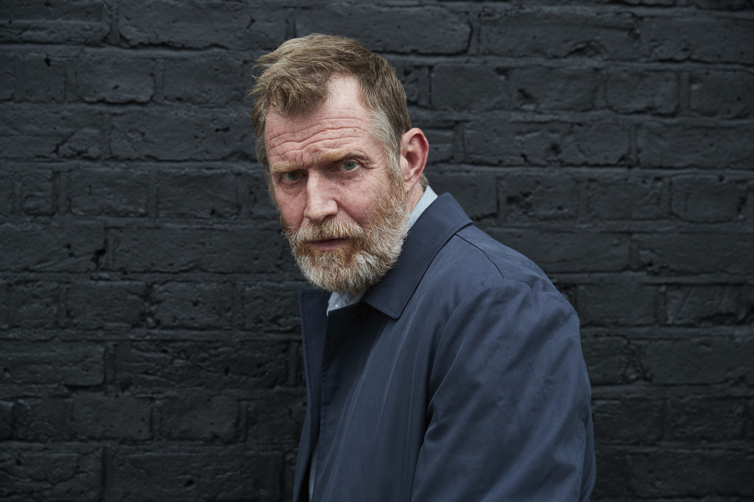 Jason Flemyng as Nick in The Walk-In, he stands against a wall and has a blue jacket on, he looks into the camera with a intense expression