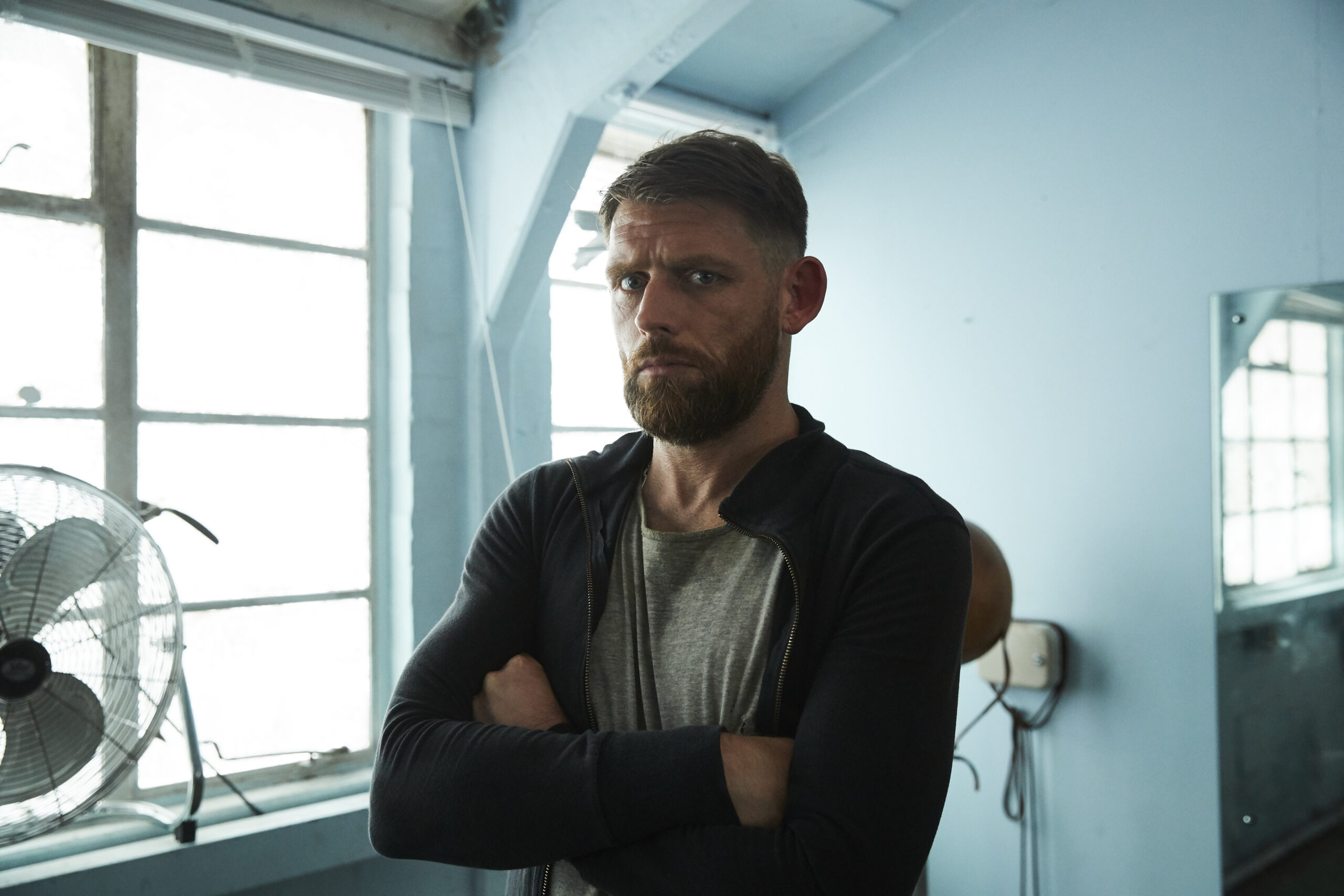 Chris Coghill as Chris in the walk-in cast, he stands with his arms folded in a white room with an angry expression