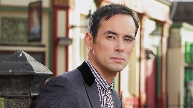 Steve John Shepherd as Michael Moon in EastEnders, he stands outside looking into the camera with a grumpy expression