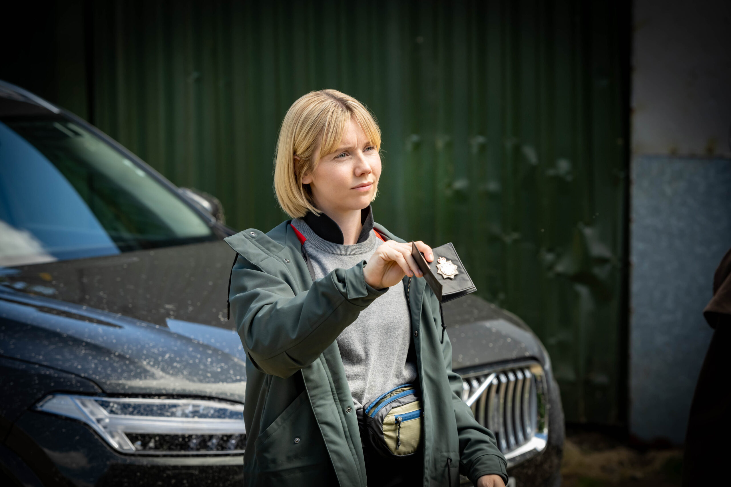 Lauren Lyle as Karen Pirie in Karen Pirie S1E1, she holds up her police badge standing outside, next to a black police car