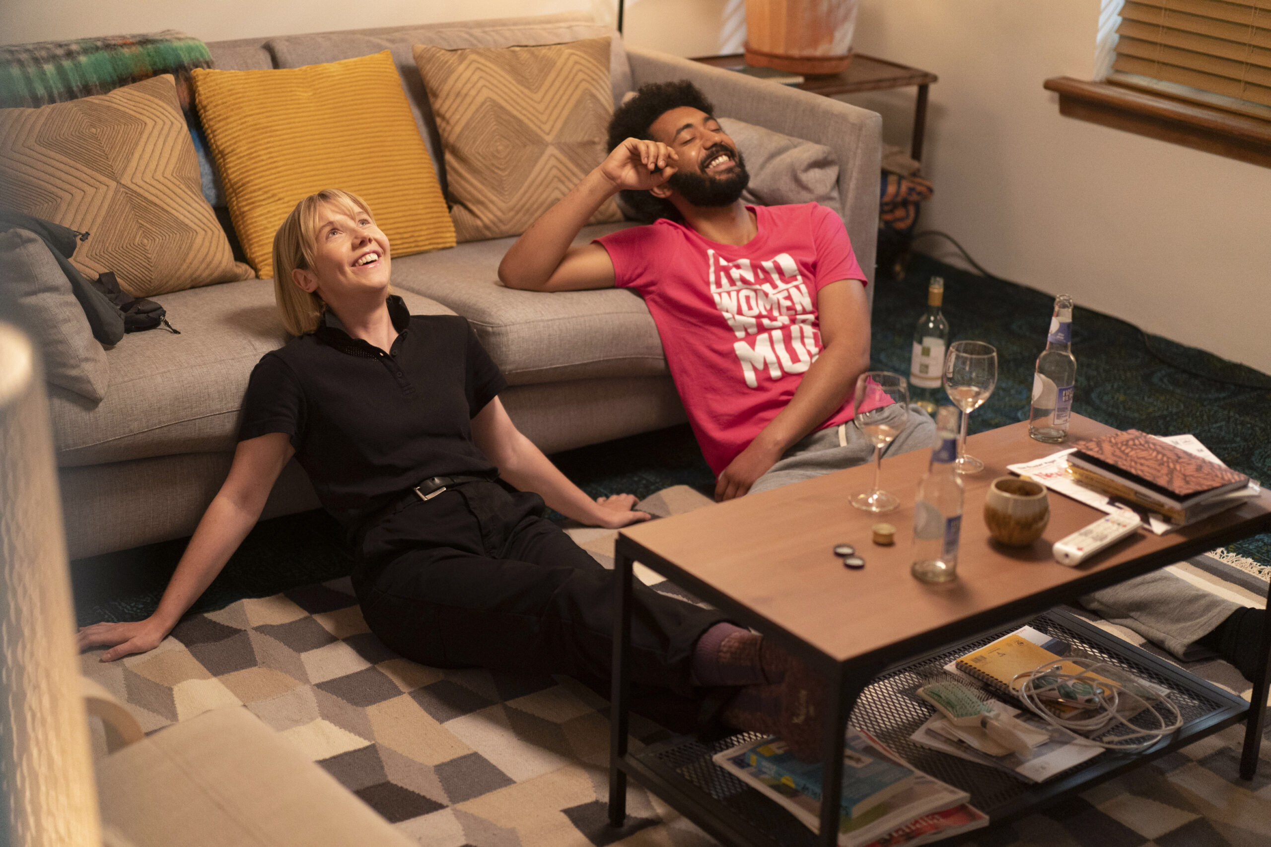 Lauren Lyle as KAREN PIRIE and ZACH WYATT as DS Phil Parhatka, they sit next to each other in a living room laughing with wine glasses in front of them