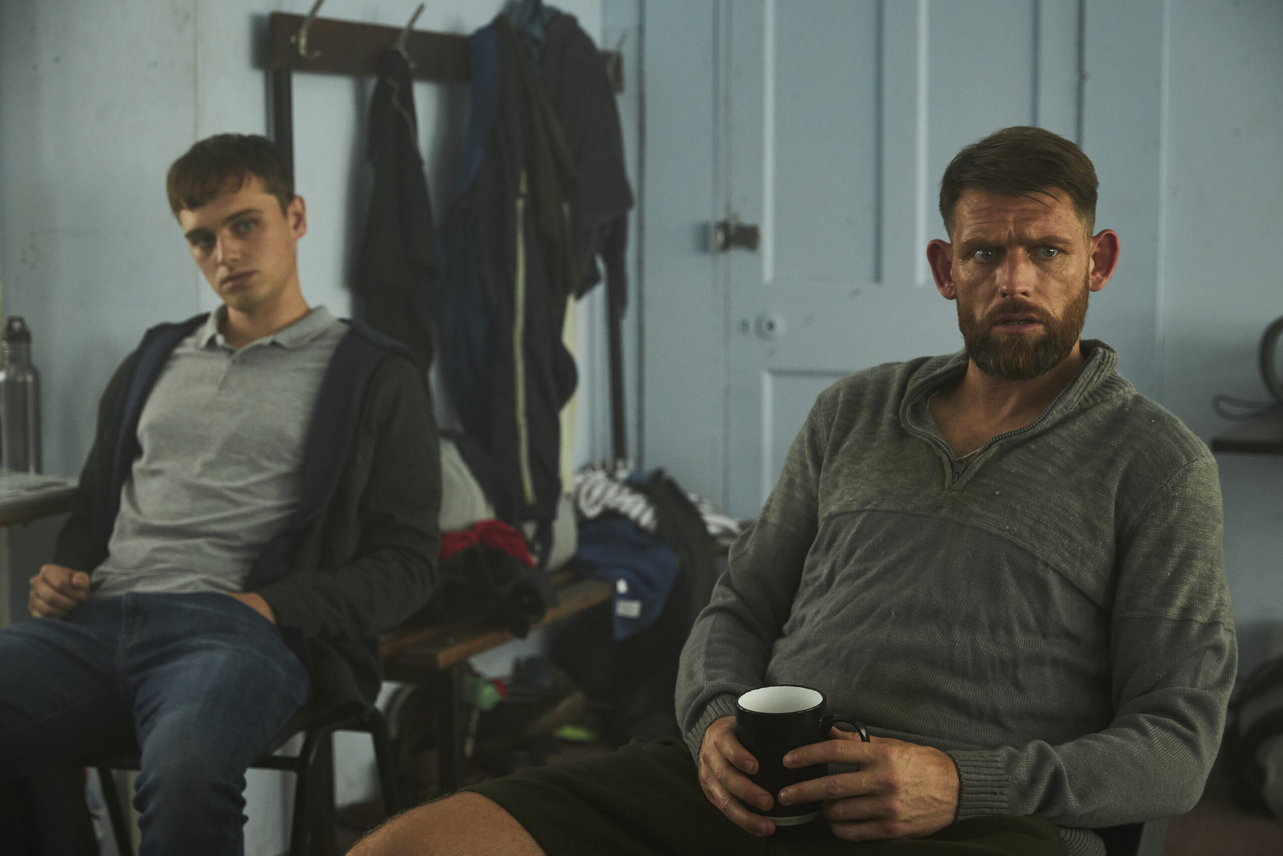 Chris Coghill as Chris Lythgoe (right) and Dean Charles Chapman as Jack (left), they sit in a room with solemn expressions with cups of coffee in their hands