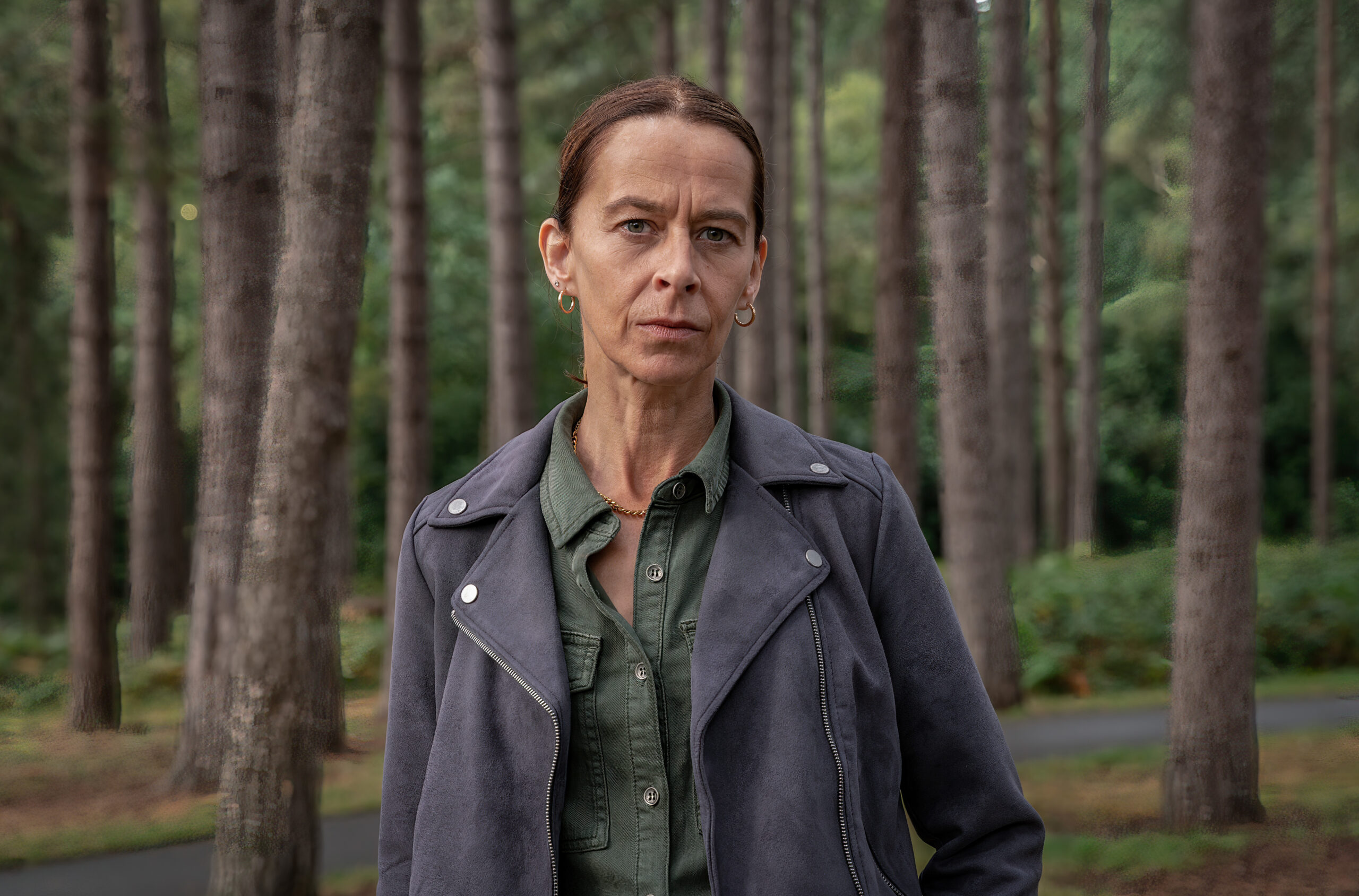 Katie Dickie as Morag in Inside Man, she stands in a forest with a small smile in a black leather jacket