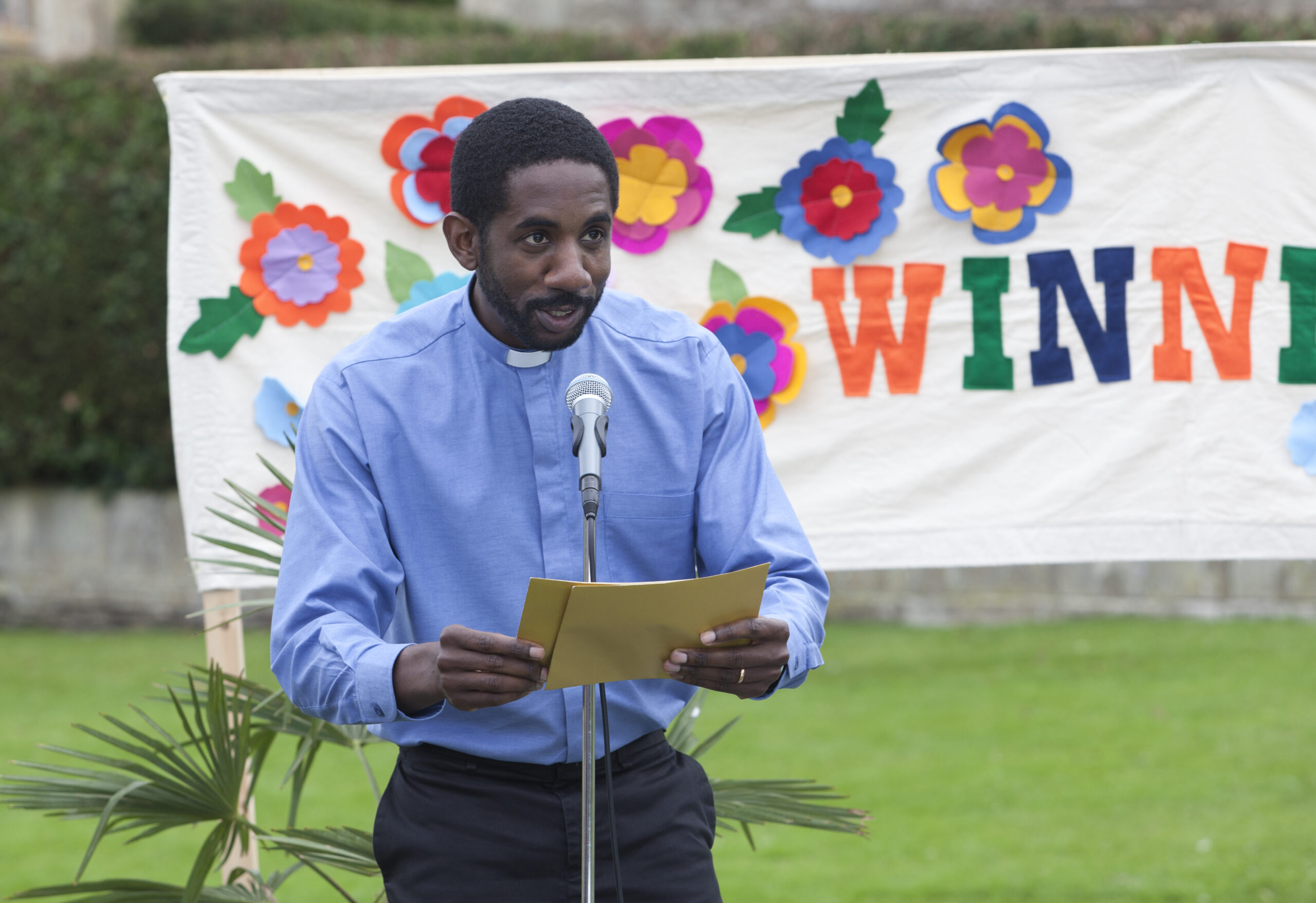 Rhashan Stone as Jez Bloxby, he stands in a suit outside in front of a sign