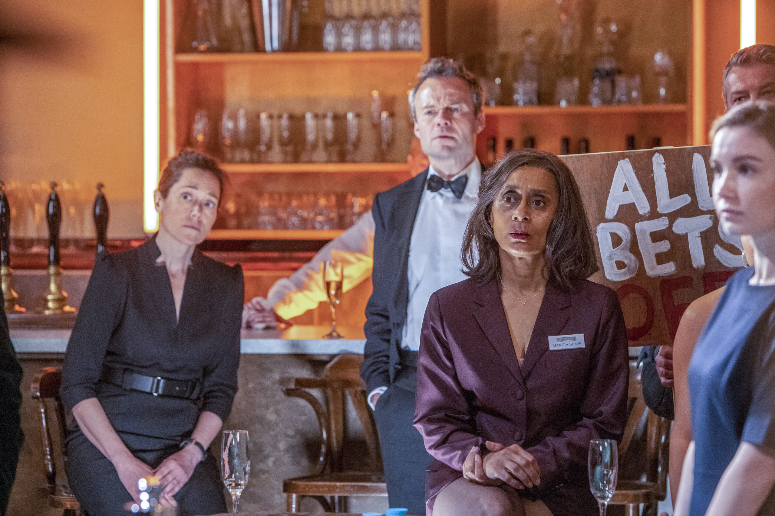 Suzanne Packer as Marcia in Agatha Raisin, she sits in a bar in a dress with a shocked expression on her face