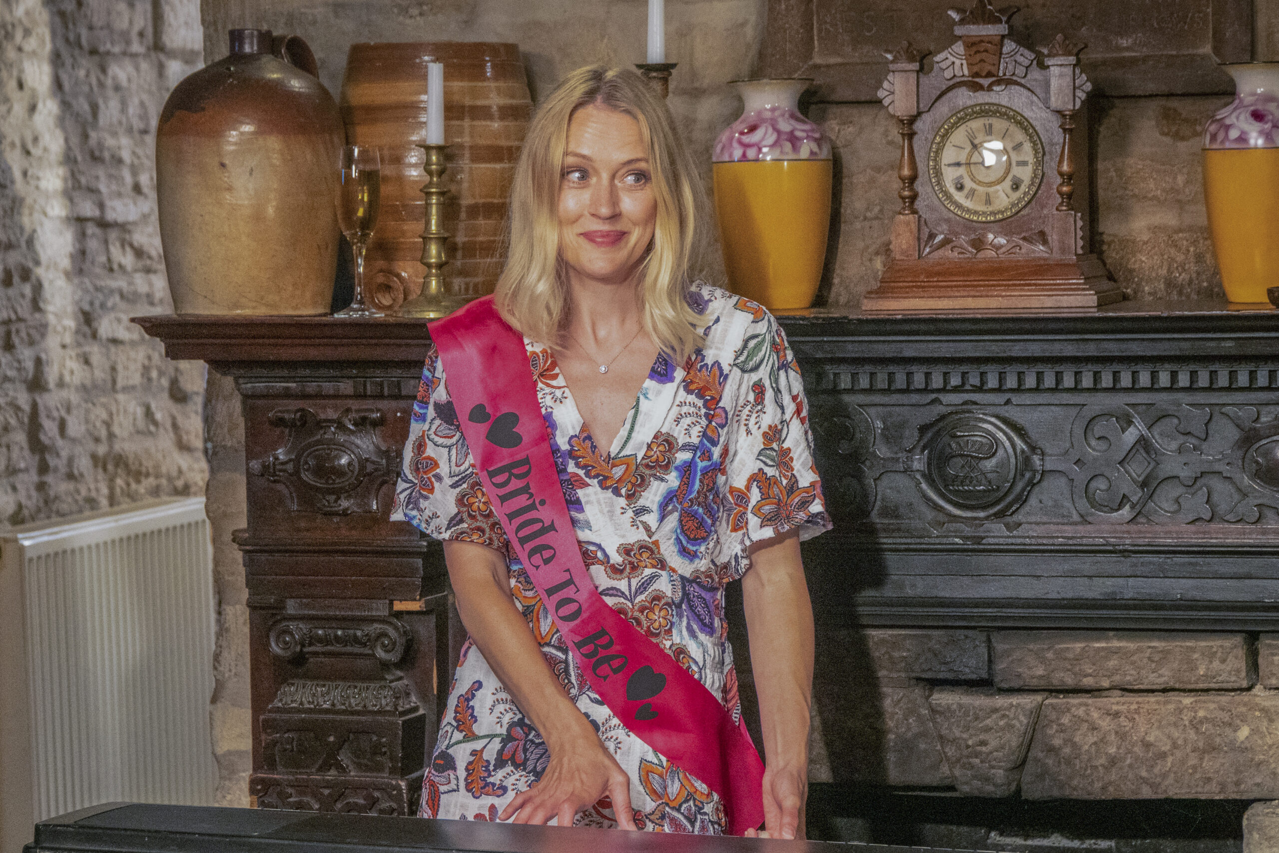Elen Rhys as Eirwen Jones in Agatha Raisin, she smiles looking to her right with a bride to be sash across her
