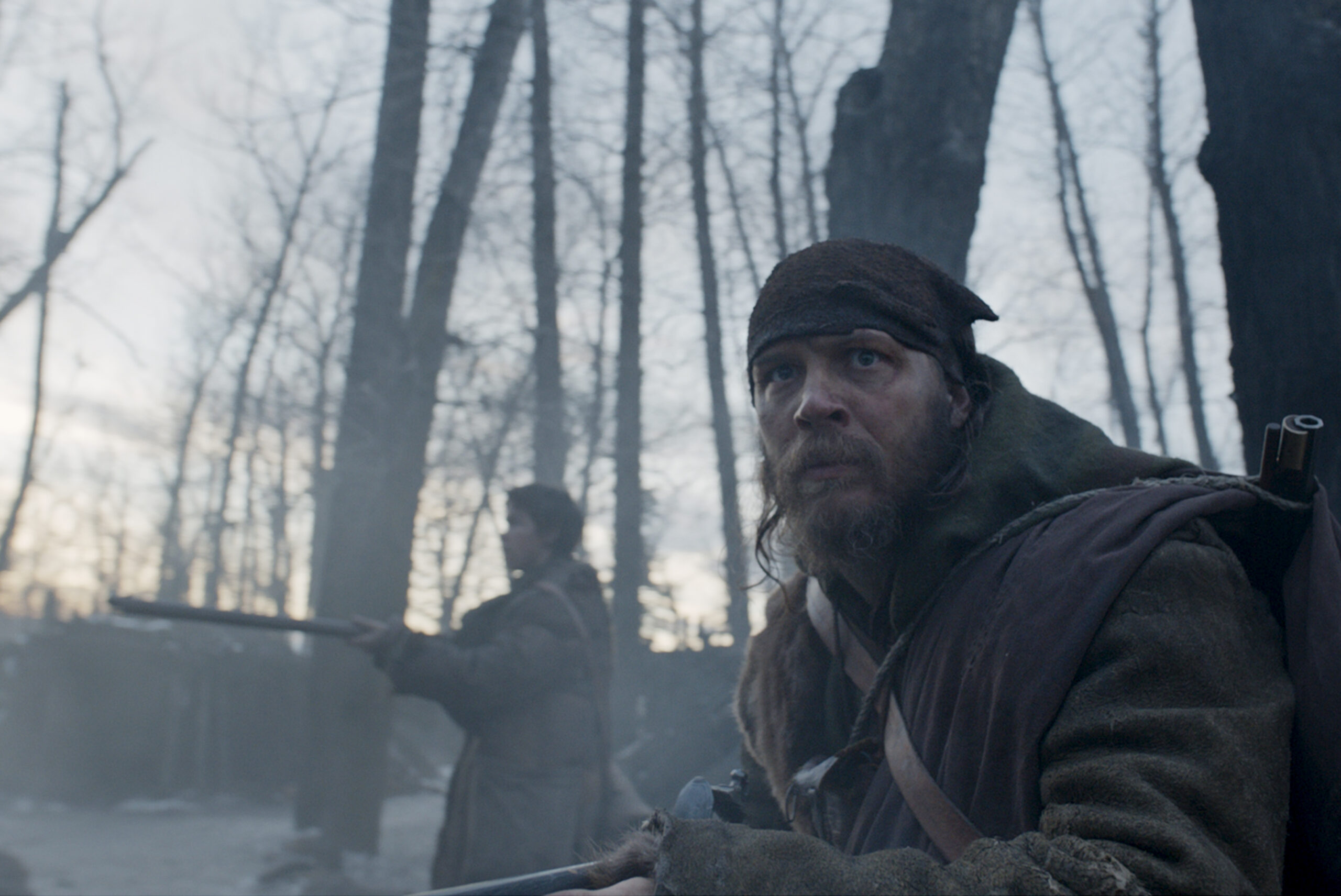(L-R): Will Poulter as Jim Bridger and Tom Hardy as John Fitzgerald in THE REVENANT. They stand in a forest, he squats down with a worried expression, holding a weapon