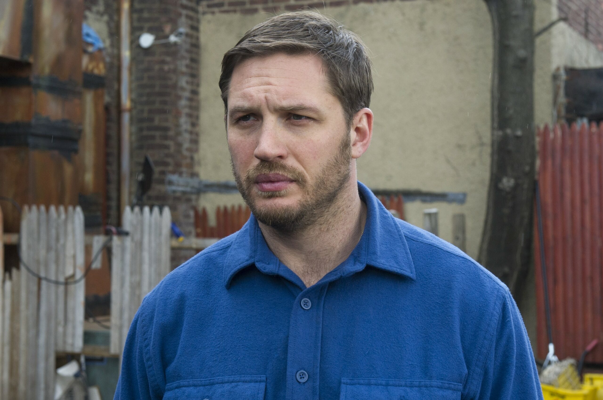 Tom Hardy as “Bob” in THE DROP. He looks to his left with a small smile, he wears a blue shirt outside