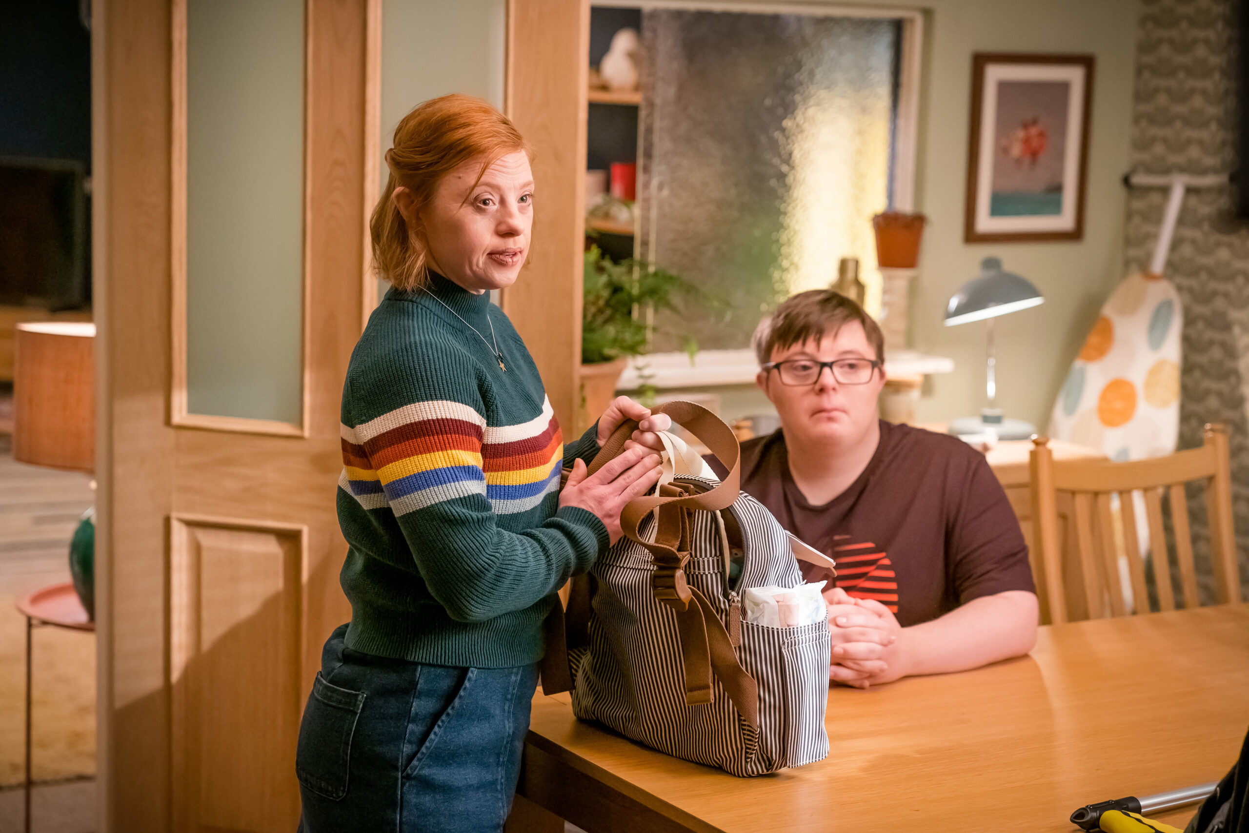 Sarah Gordy as Katie (left) and Leon Harrop as Leon (right), they sit at a table as they look at someone with neutral facial expressions