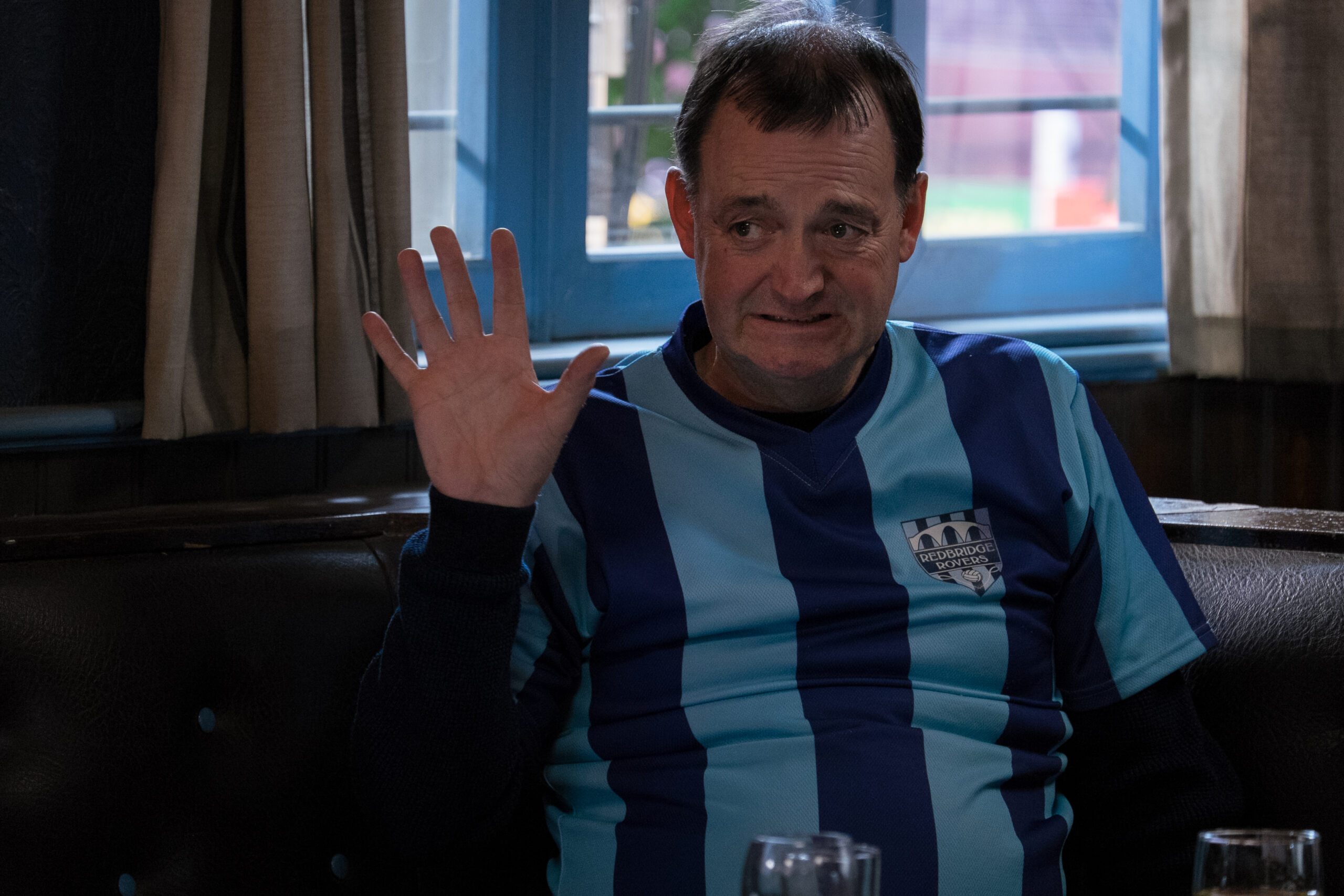 Craig Cash as Pete in Rovers, he waves at someone to his left with an awkward smile on his face and a football top on