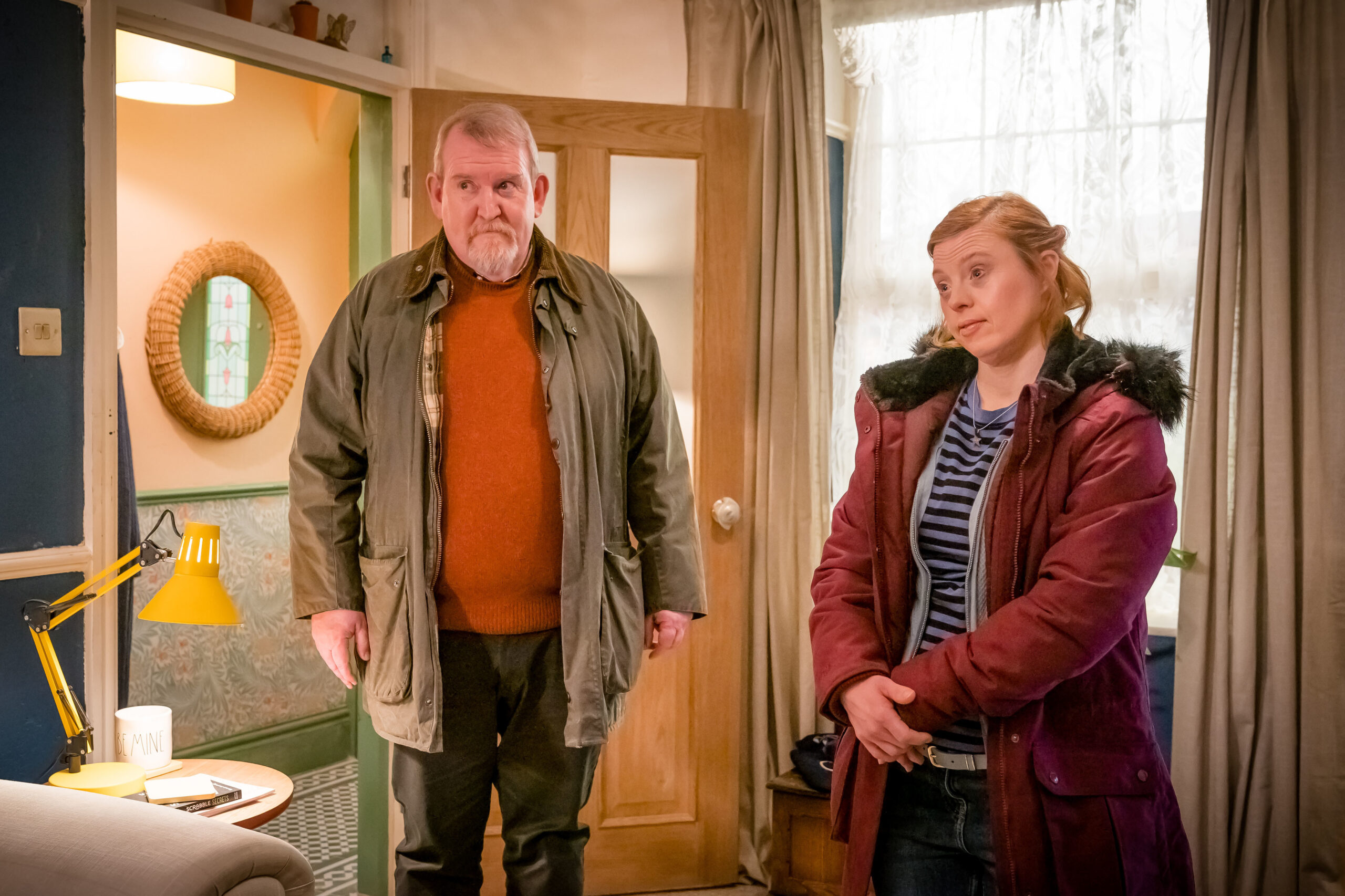 Nigel Betts as Steve Thorne in Ralph & Katie, he stands next to Katie in her home with a small smile on his face and his coat on