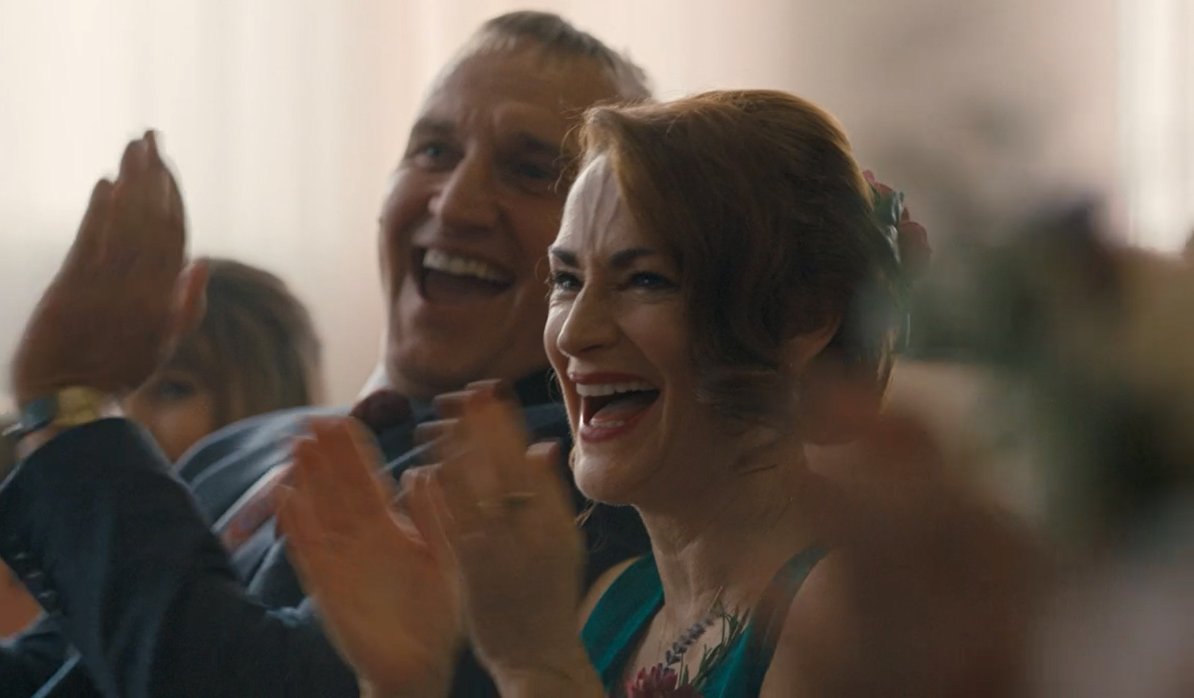 Pooky Quesnel as Louise sit next to Christopher Ecclestone as Maurice in Ralph & Katie's wedding in The A Word, they clap and smile