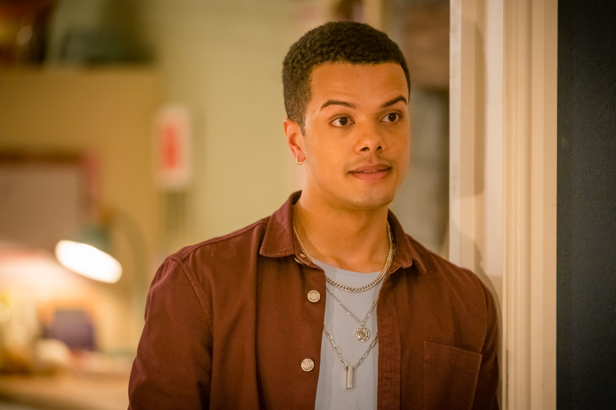 Dylan Brady as Danny in Ralph & Katie, he stands in a living room with a small smile with a brown jacket on