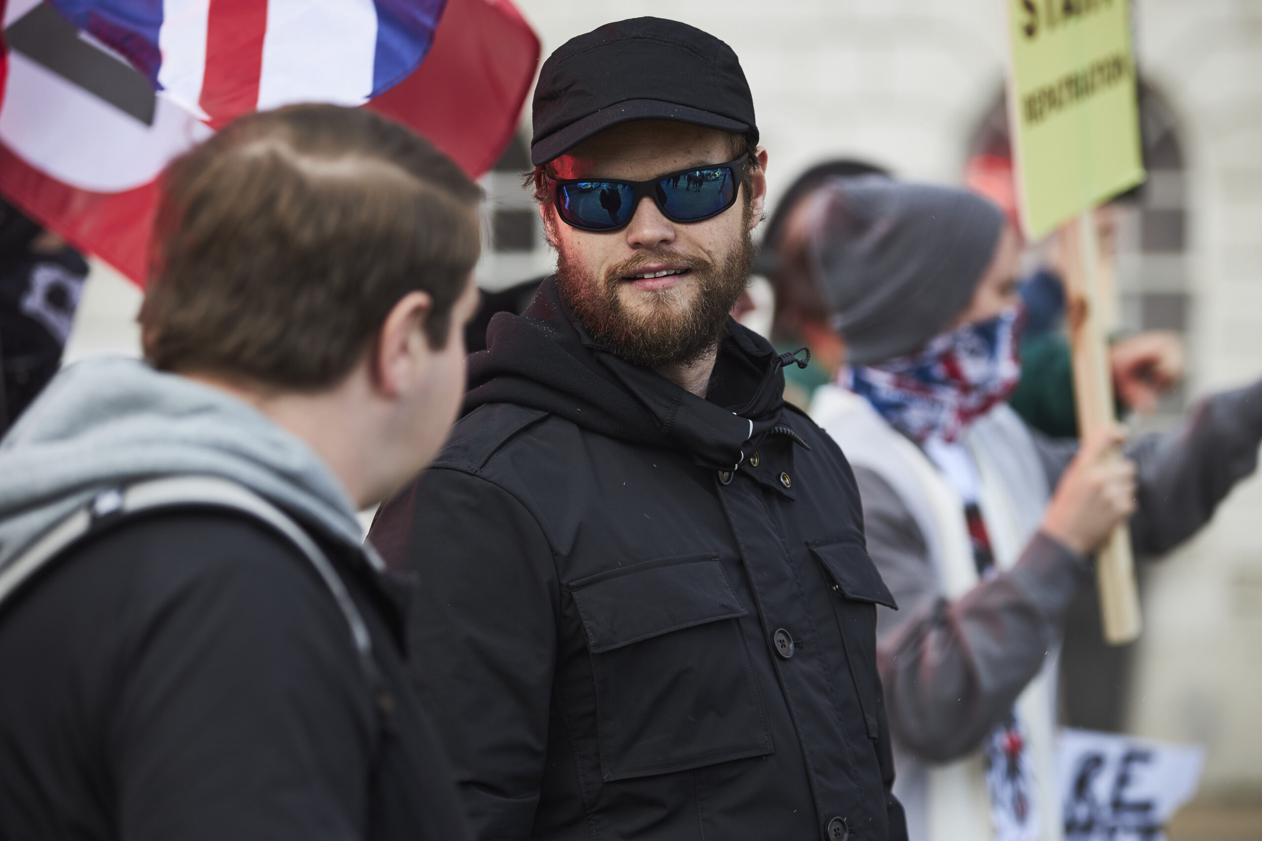 Bobby Schofield at Matt in The Walk-In, he stands at a protest in dark clothes and dark sunglasses