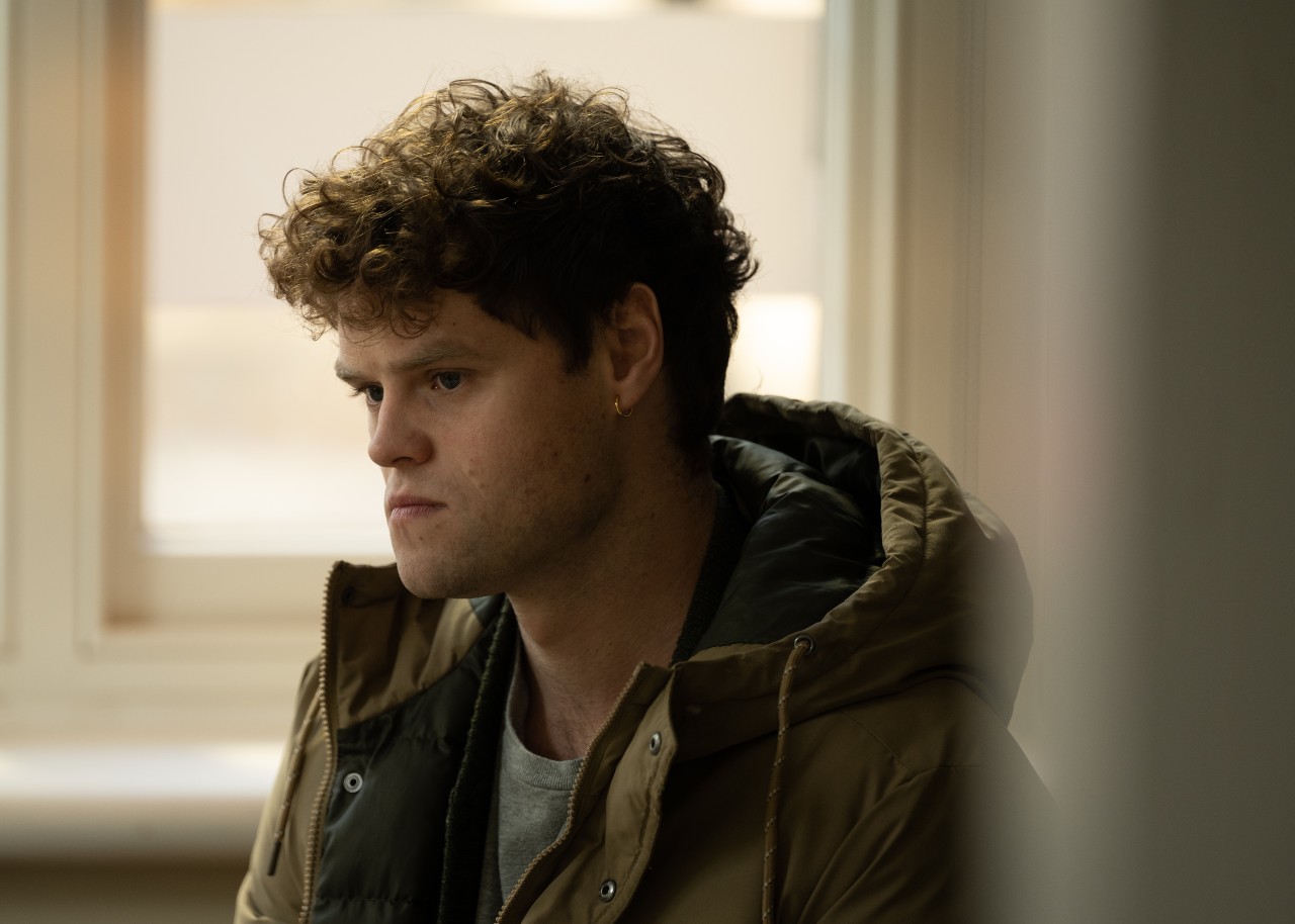 Bobby Schofield as Bobby Moran in The Suspect, he looks to the side with a grumpy face in a big coat